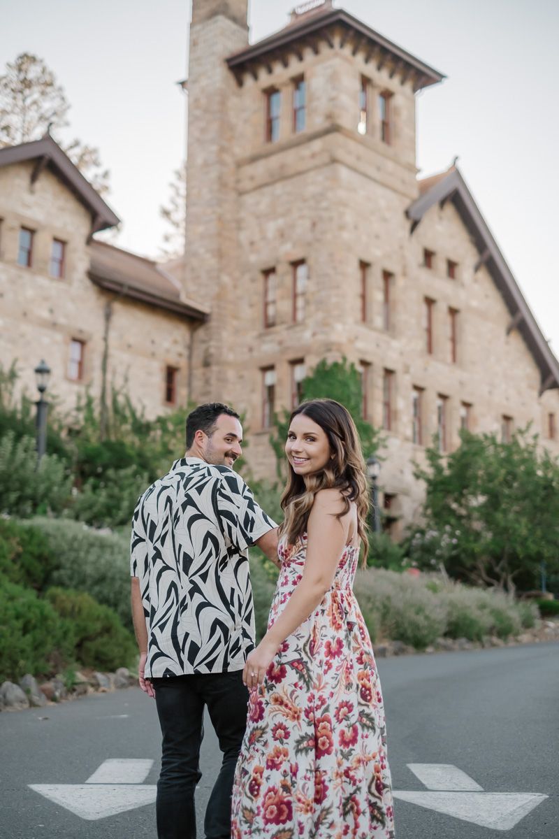 Couple holding hands in front of a stone castle building, woman in floral dress and man in black and white shirt.