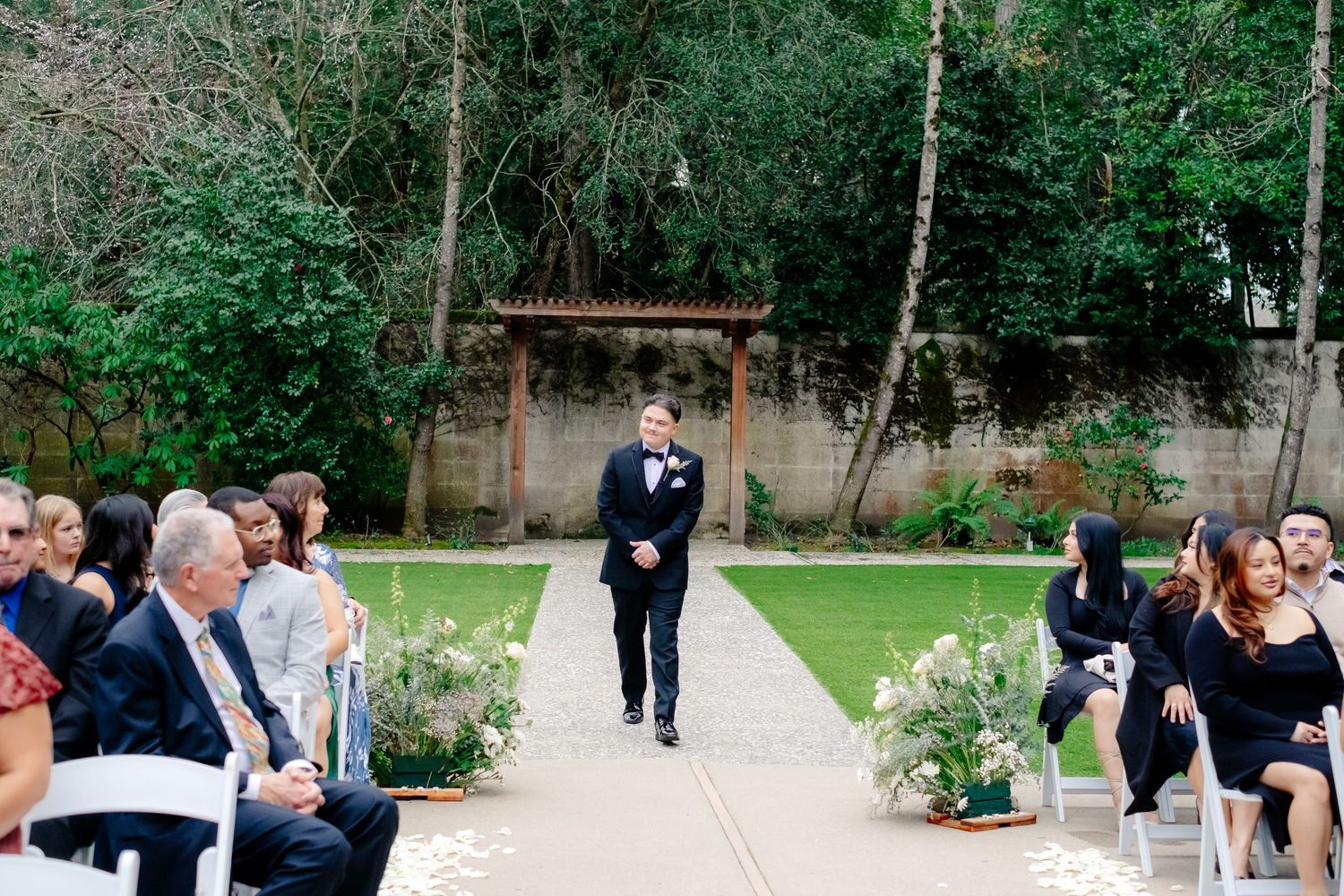 Groom walks down outdoor wedding aisle flanked by seated guests and floral arrangements at garden ceremony.
