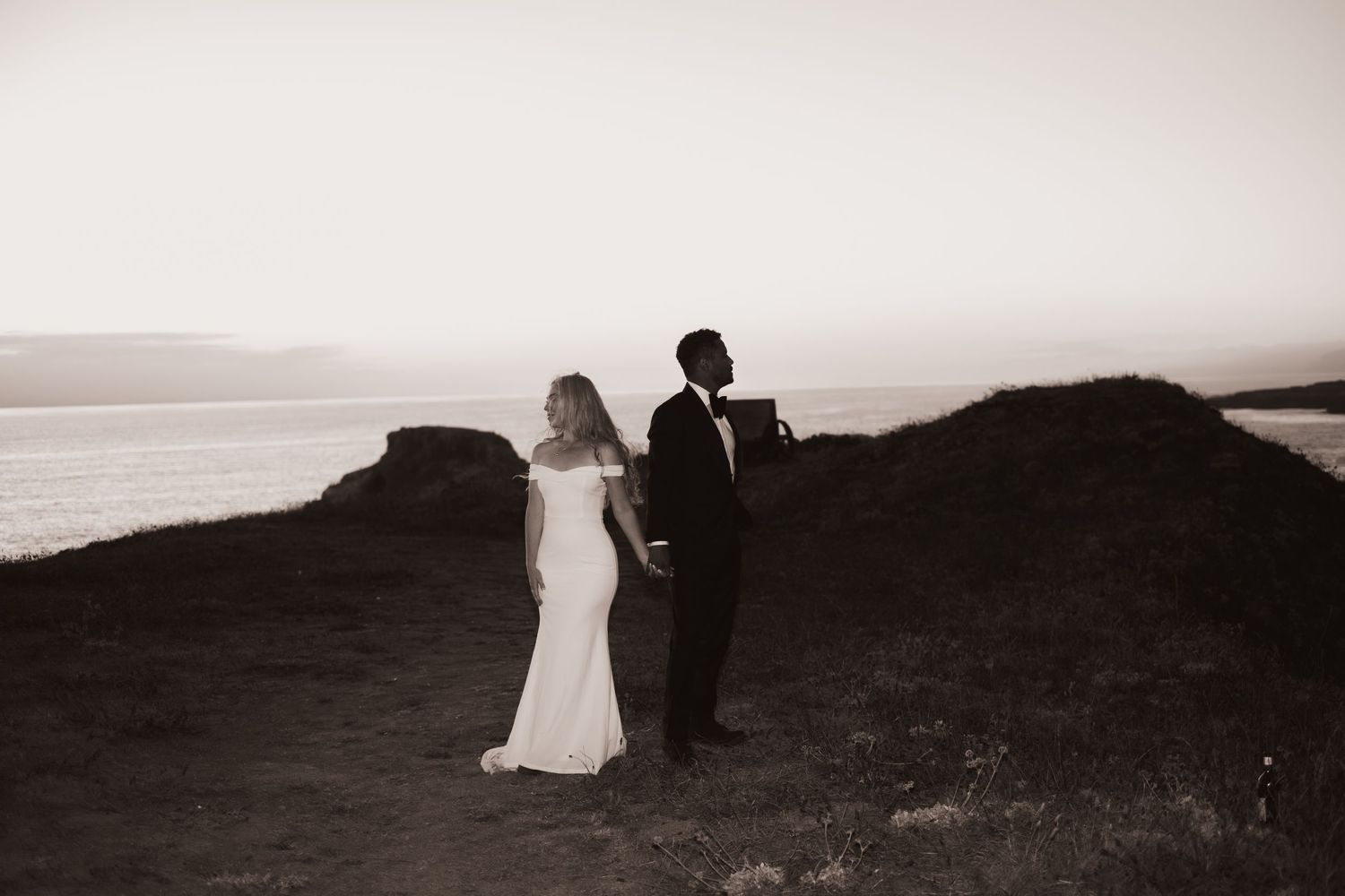 Black and white silhouette of bride and groom standing on coastal cliffs at sunset.