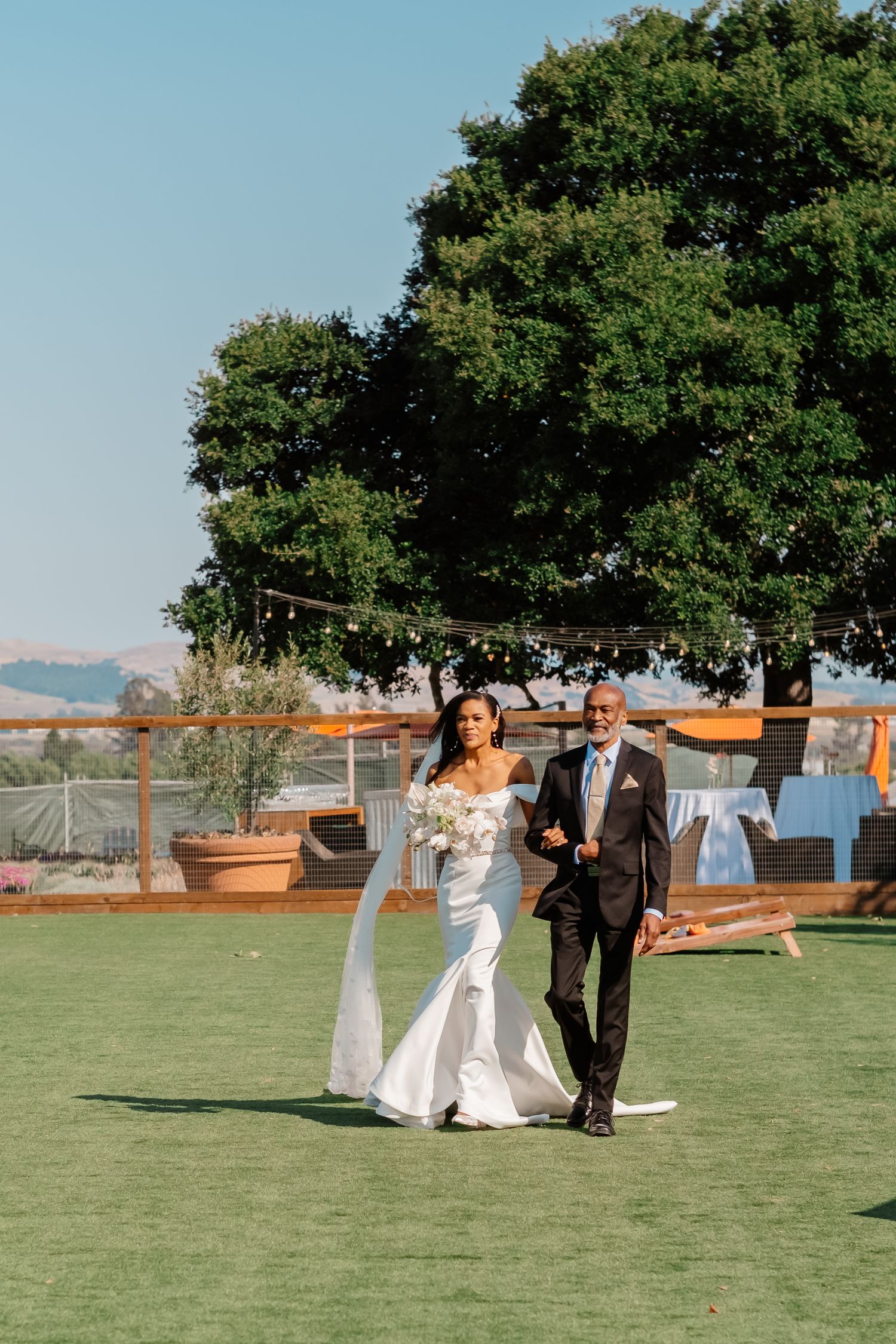 Bride in white off-shoulder gown walks with suited man across green lawn at outdoor wedding venue.