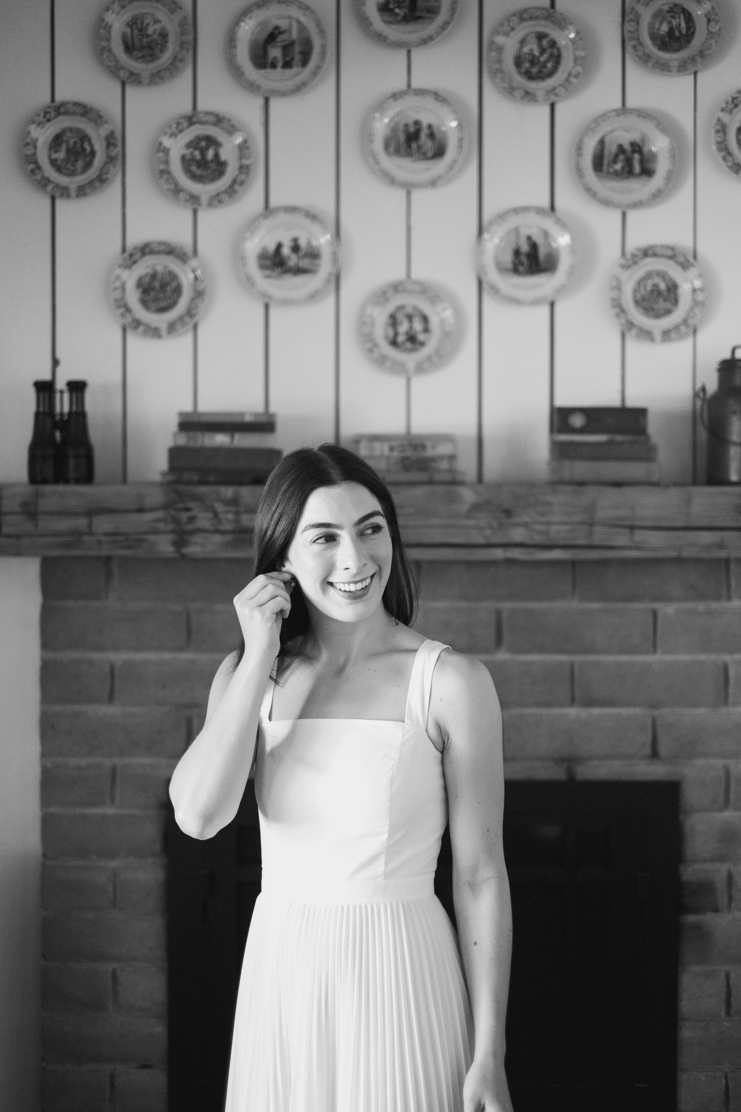 Smiling bride in white pleated dress posing before decorative plates above fireplace.