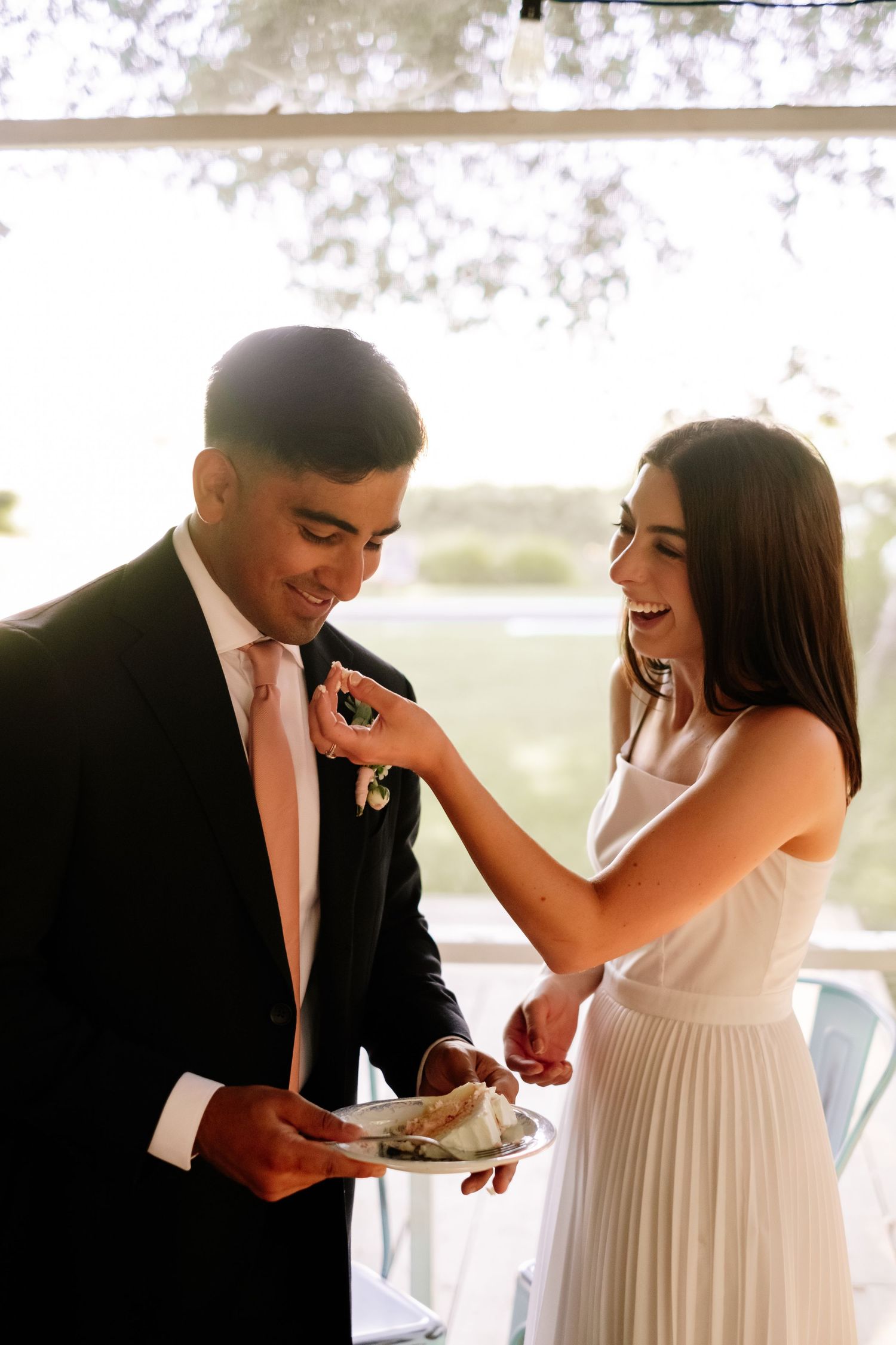 Laughing bride feeding wedding cake to groom in navy suit during sunny outdoor reception.