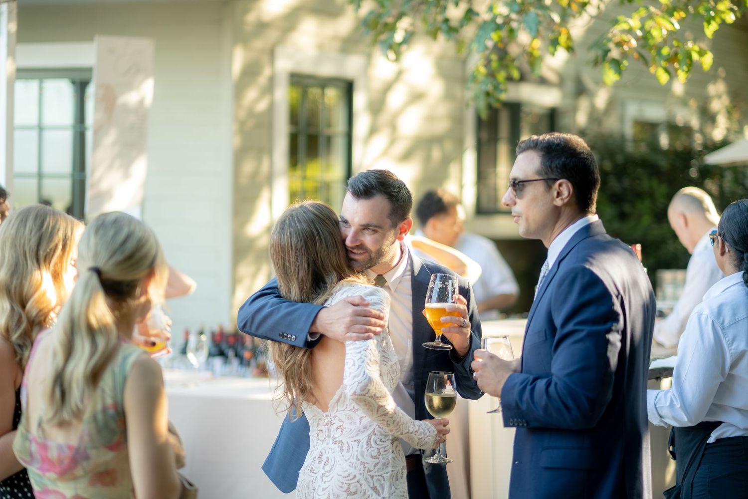 Bride in lace gown embracing a guest at a sunny outdoor wedding cocktail reception.