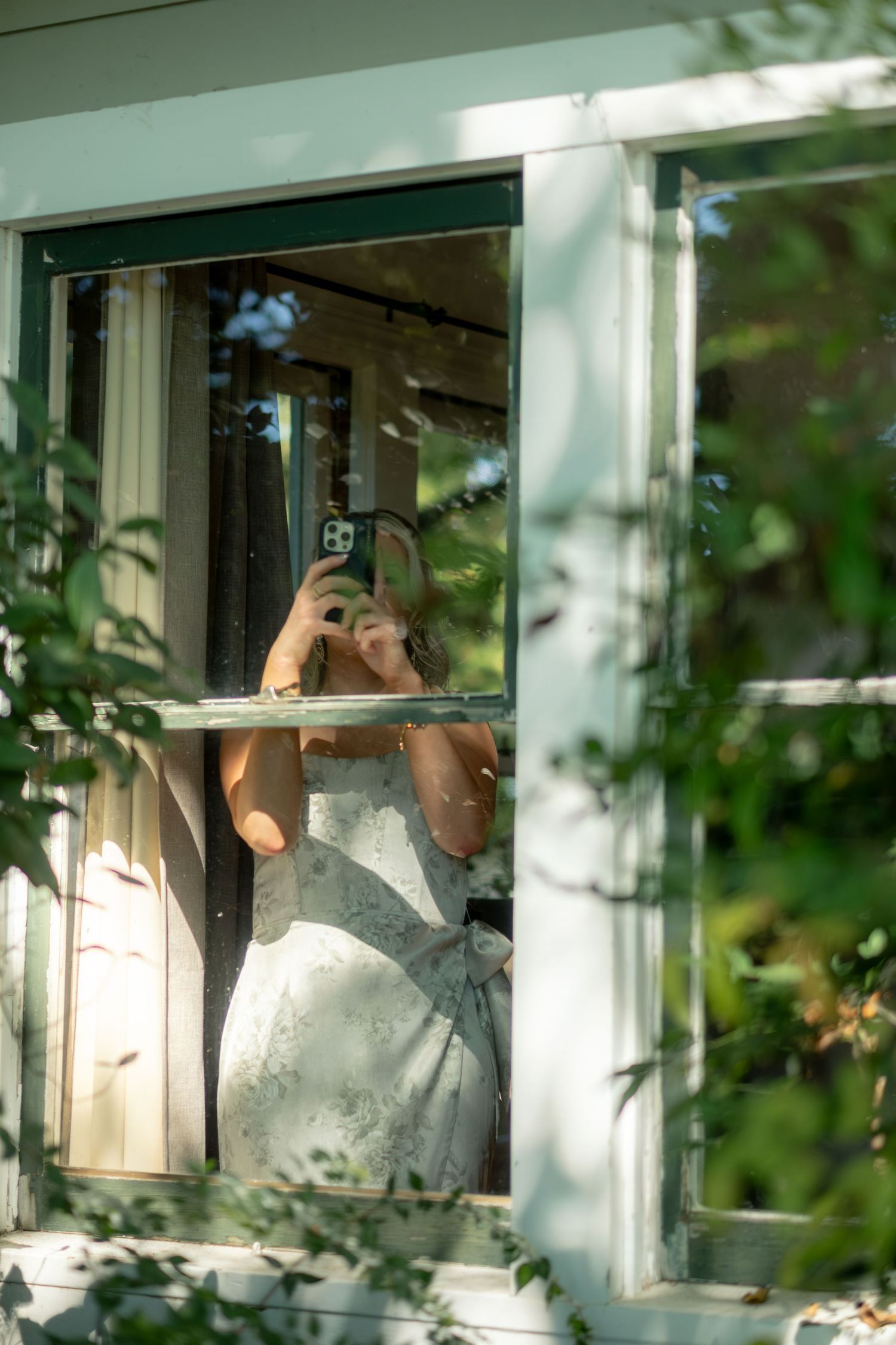 Bride in white wedding dress taking a mirror selfie through an open window surrounded by green foliage.