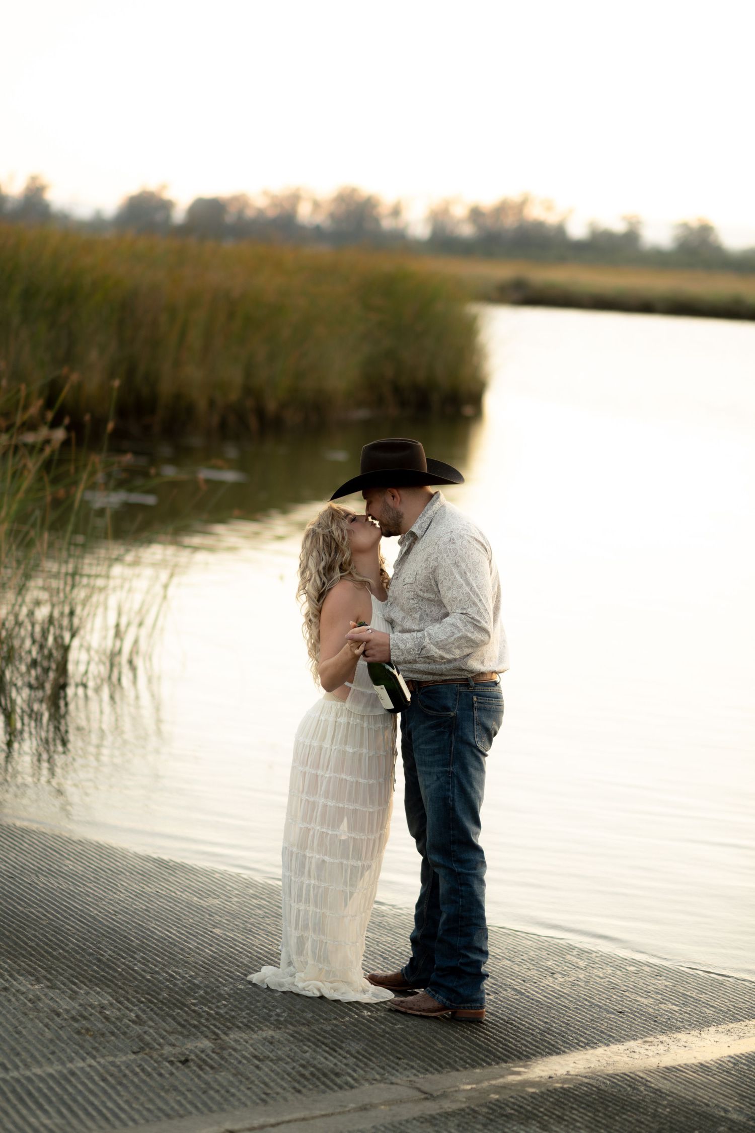 Couple sharing a kiss on a dock at sunset, man in cowboy hat and woman in flowing white dress.