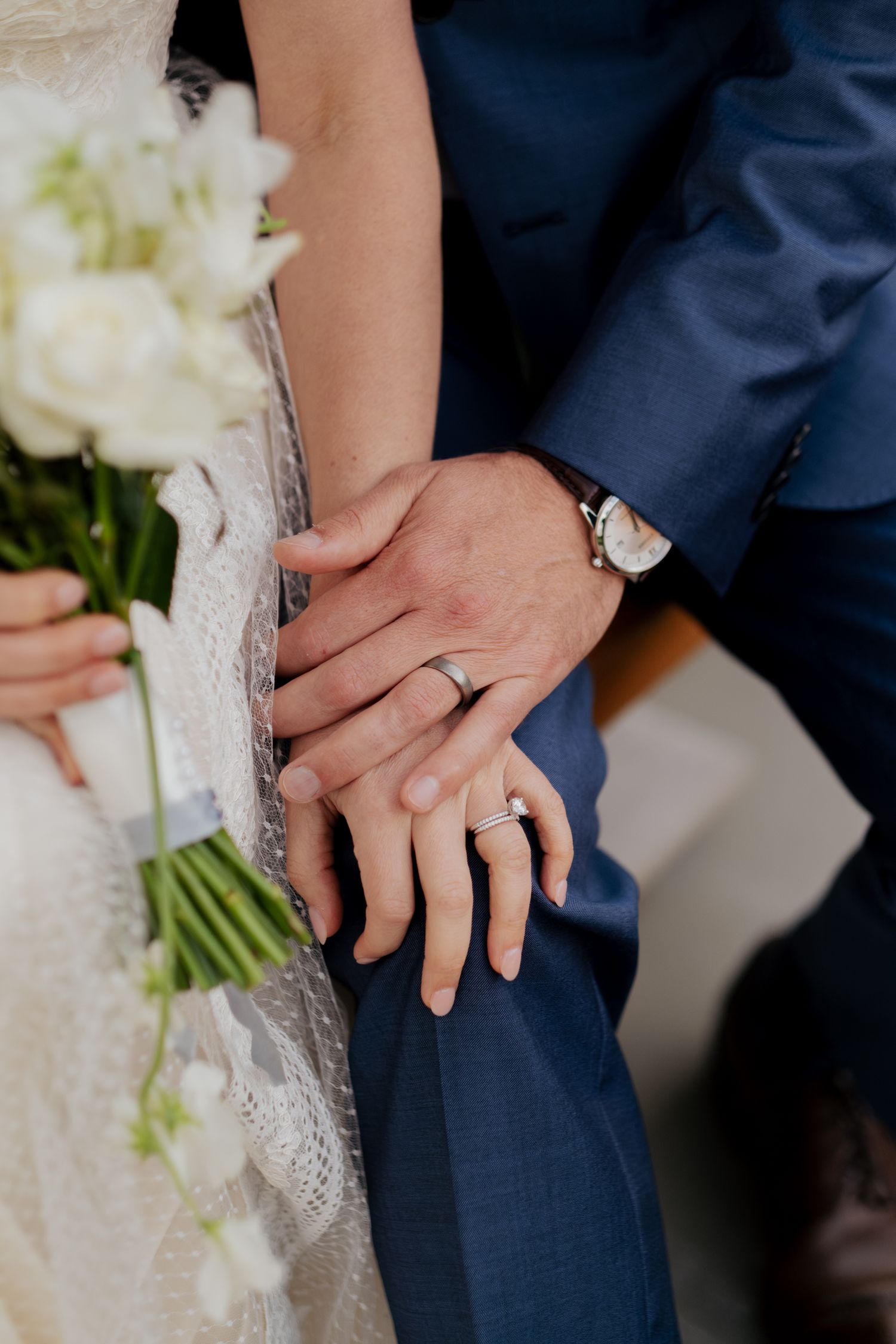 Close-up of newlyweds hands showing wedding rings, bride holding white bouquet, groom in navy suit.