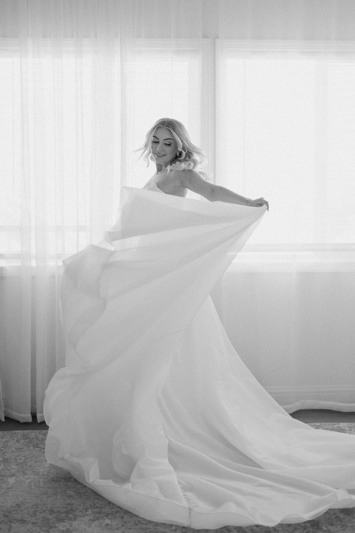 Black and white photo of a bride twirling her flowing white ball gown skirt by sheer curtains.