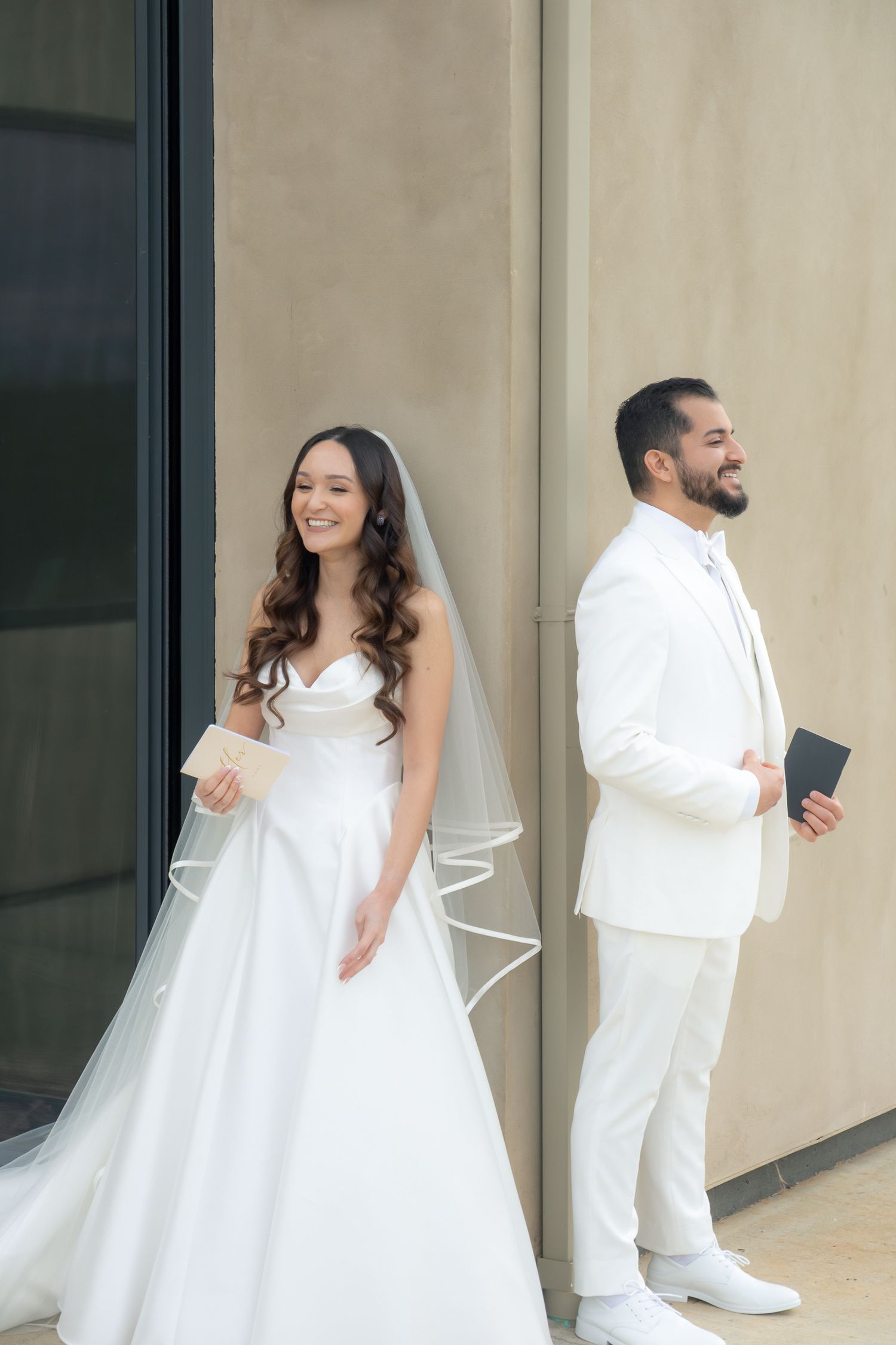 Bride in white gown and groom in white suit smile back-to-back before their first look ceremony.