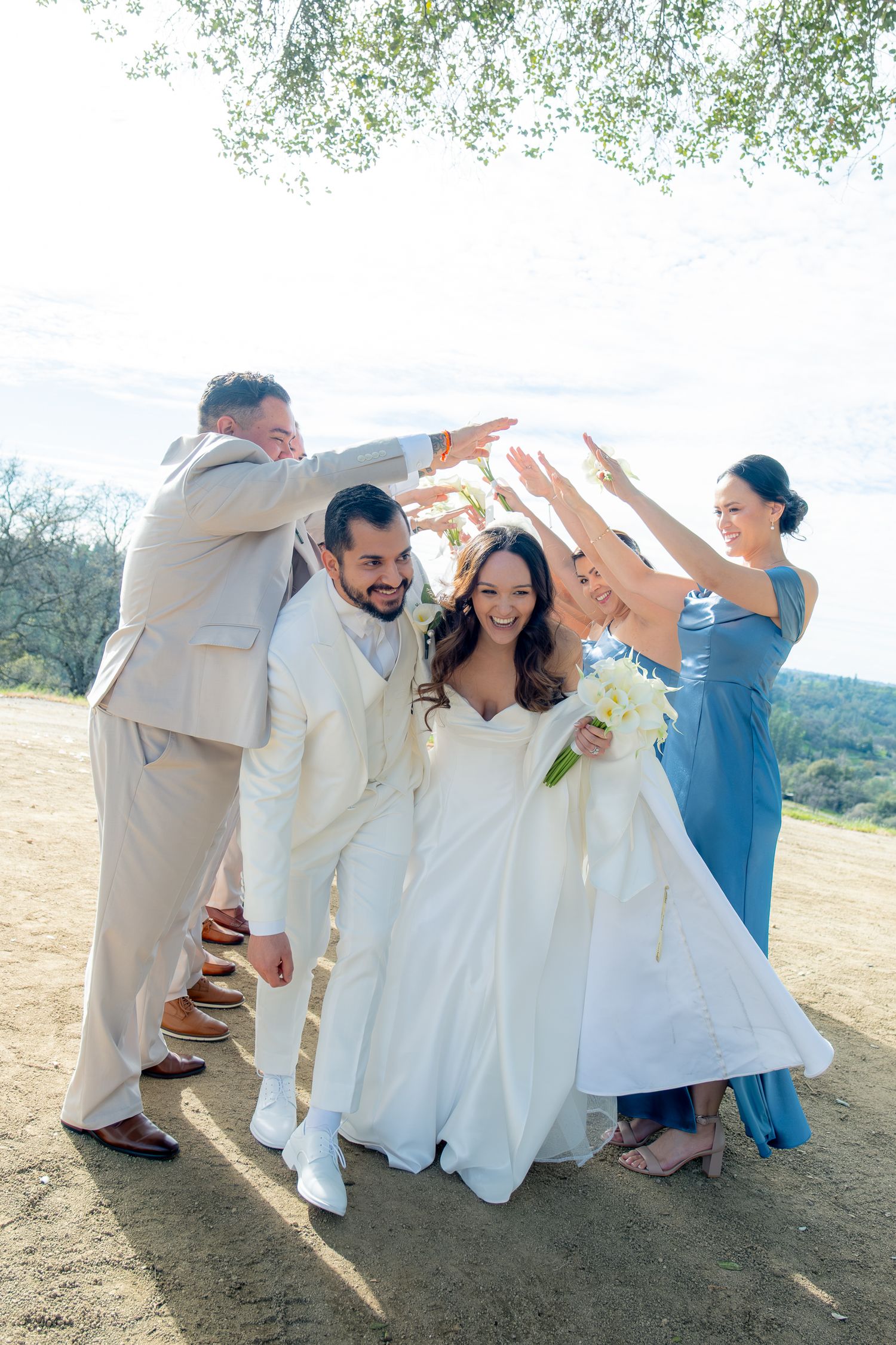 Bride and groom celebrate with wedding party raising hands outdoors under a tree at sunset.