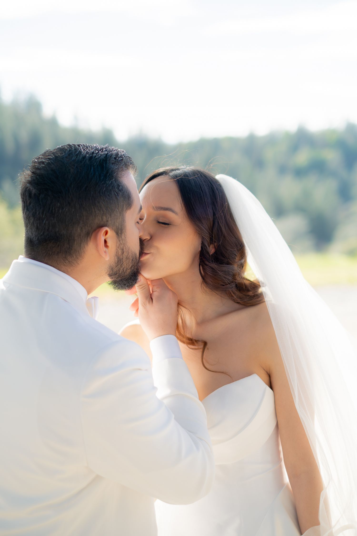 A bride and groom share a romantic kiss outdoors, dressed in white wedding attire with a lush green forest backdrop.