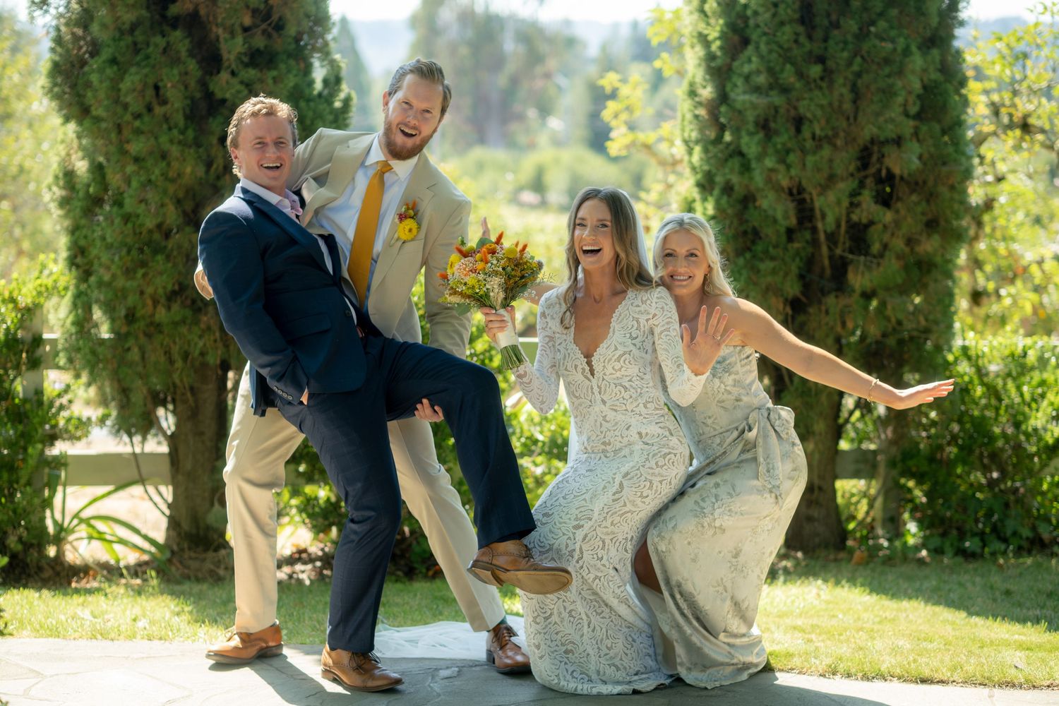 Four joyful wedding guests pose playfully outdoors, with cypress trees and sunny skies in the background.