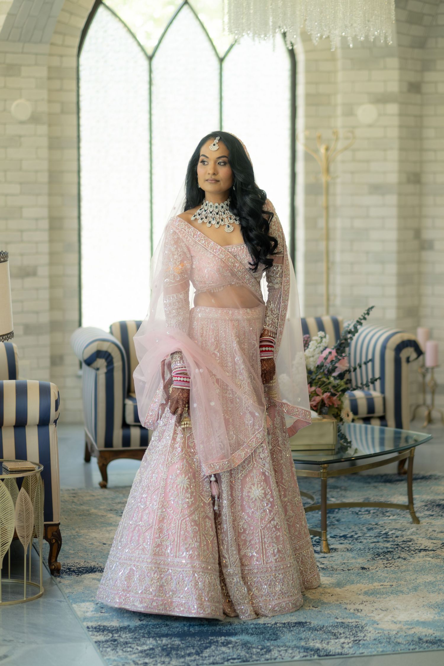 Bride in pink embroidered lehenga standing in elegant venue with arched windows and blue chairs.