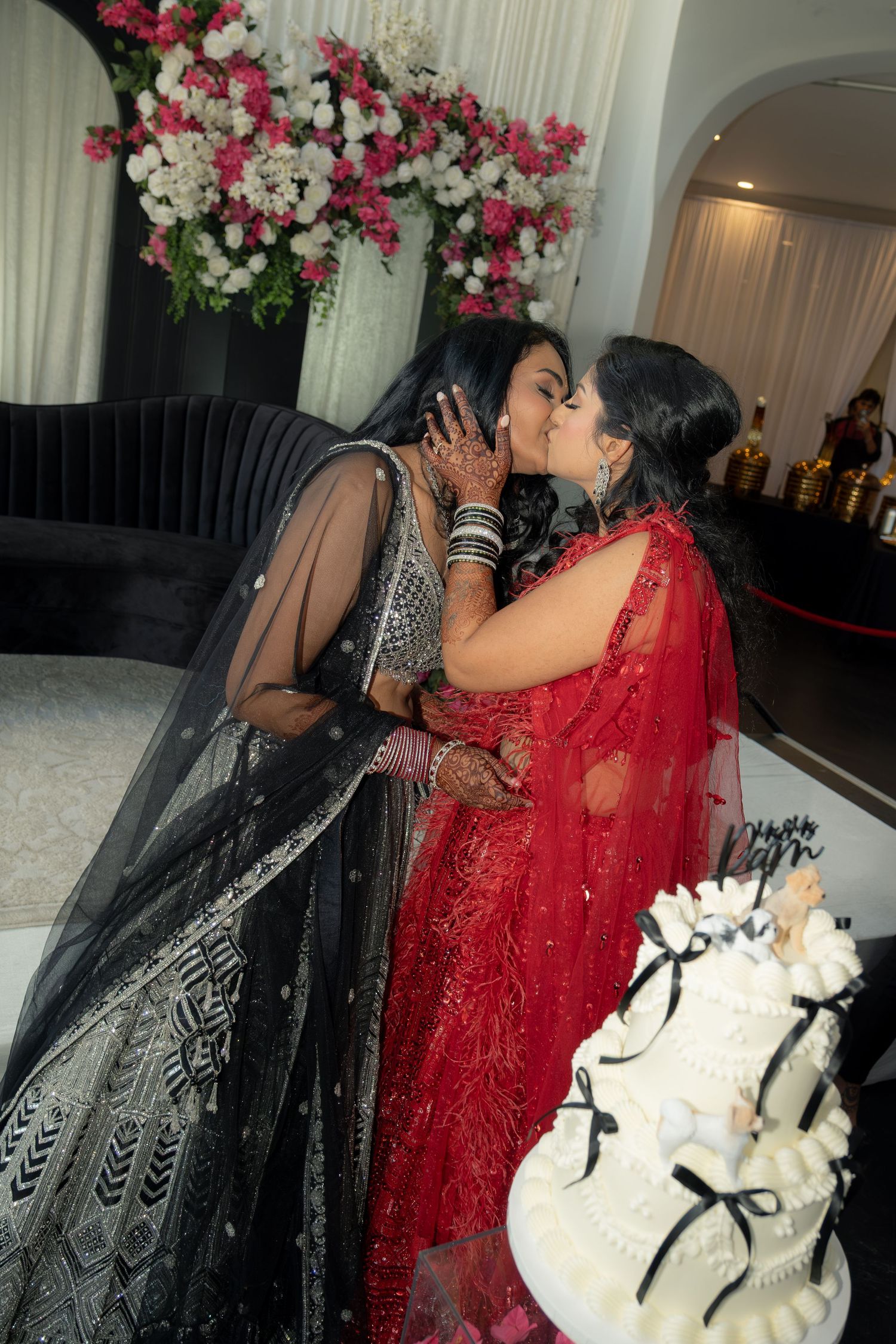 Two brides in black and red lehenga share a kiss beside a wedding cake at their Indian wedding reception.