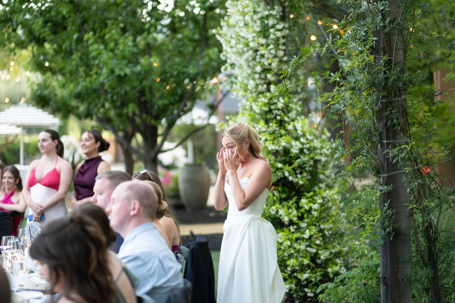 Emotional bride covering her mouth with hands during outdoor wedding reception surrounded by guests.