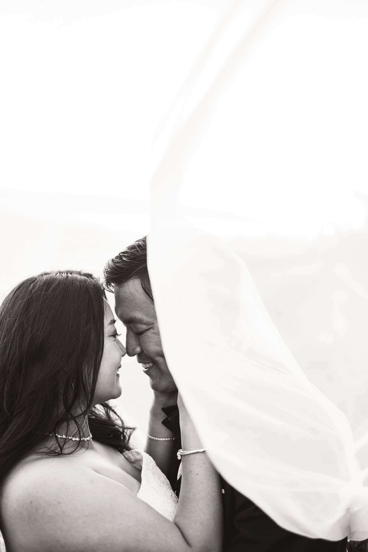Romantic black and white portrait of bride and groom nearly kissing beneath flowing wedding veil.