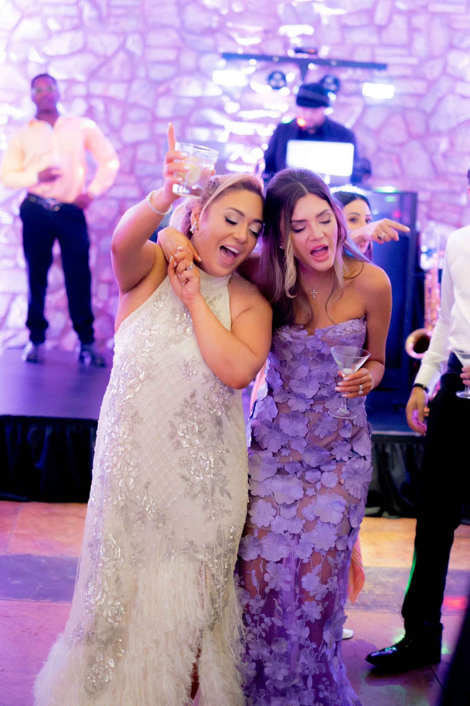 Two women in elegant gowns dancing and singing joyfully at a wedding reception with purple lighting.