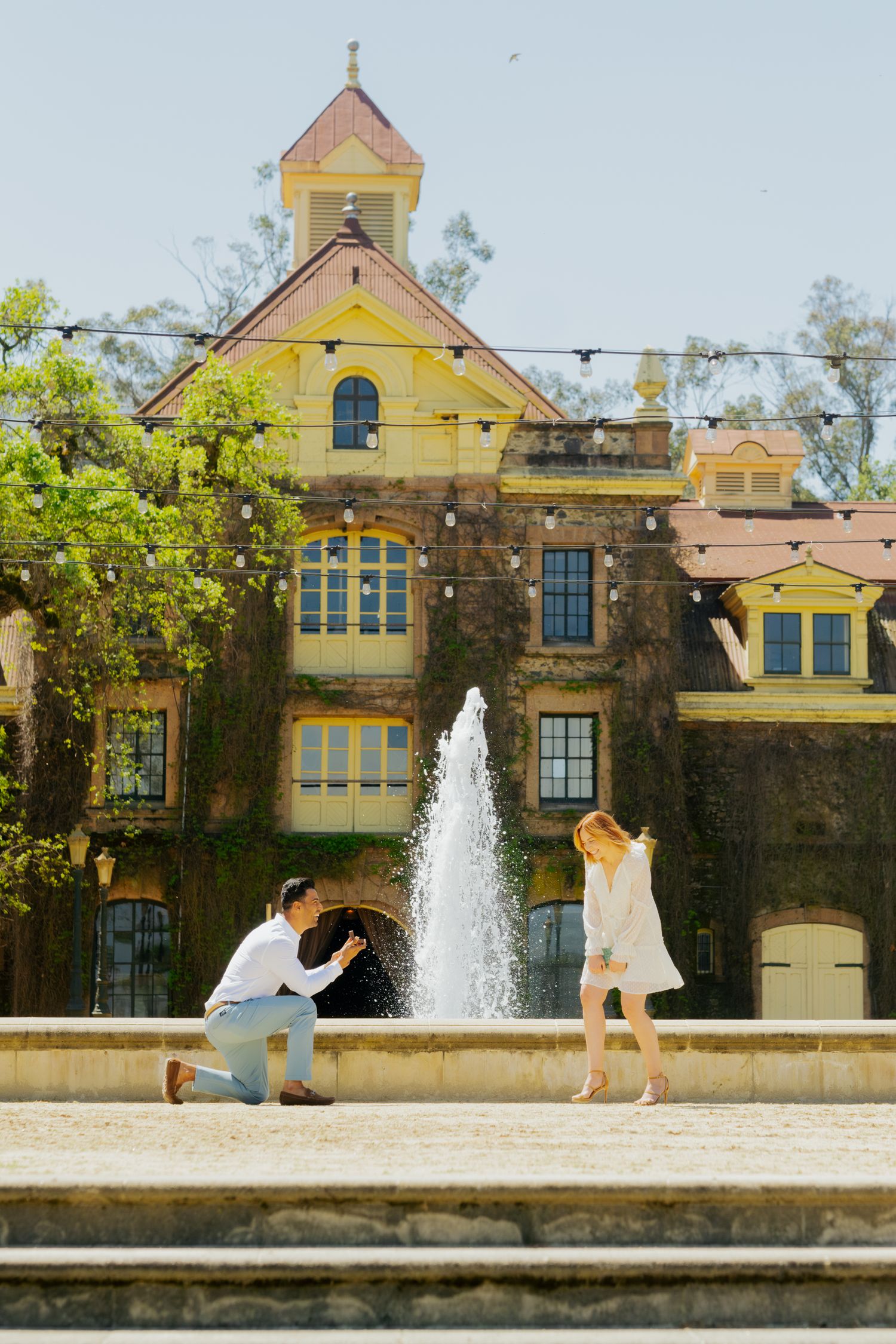 Man on one knee proposing to a woman near a fountain outside a historic yellow estate.