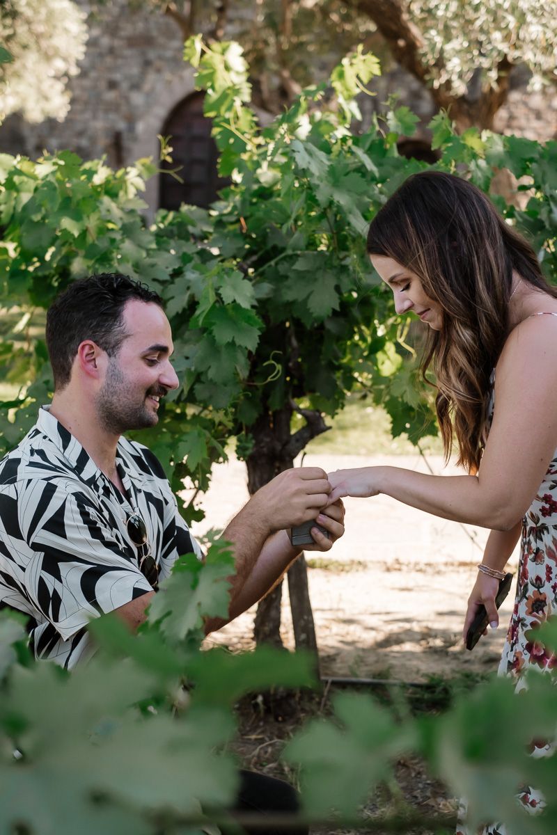 Man on one knee placing engagement ring on womans hand amid green vineyard grapevines.
