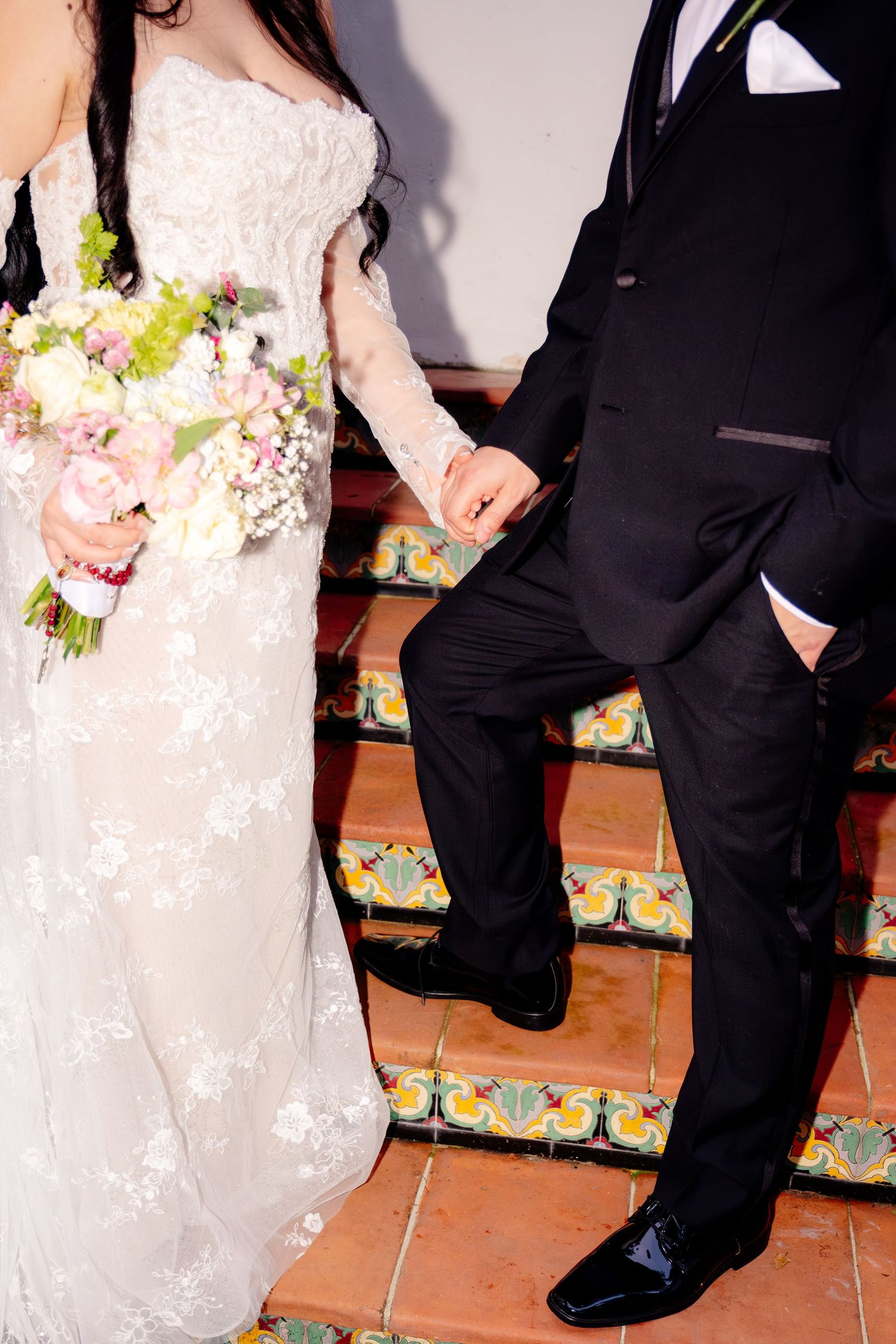 Bride holding colorful bouquet in white lace gown holding hands with groom on decorative tiled steps.