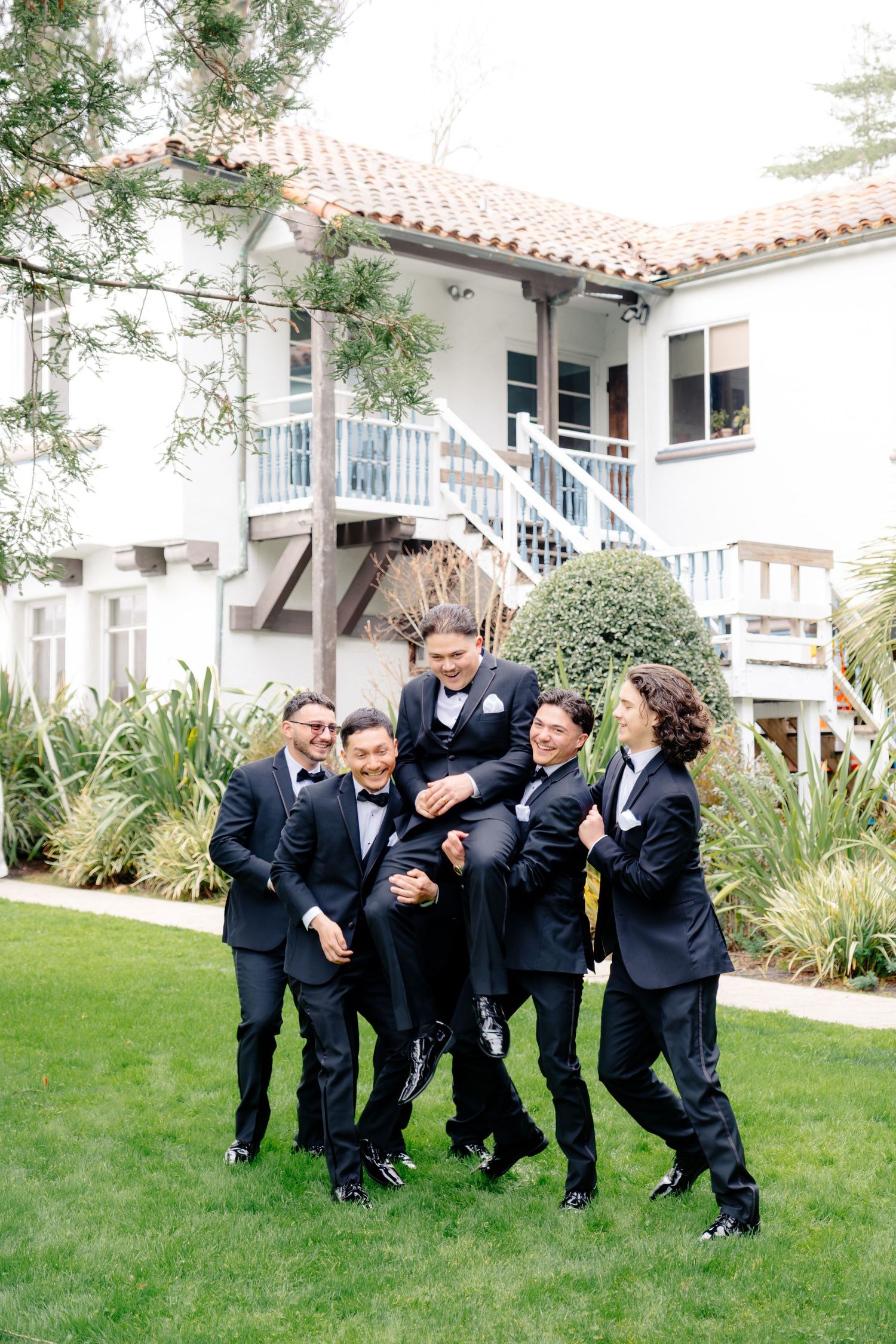 Groomsmen in black tuxedos lifting the groom outdoors on green lawn in front of white Spanish-style venue.