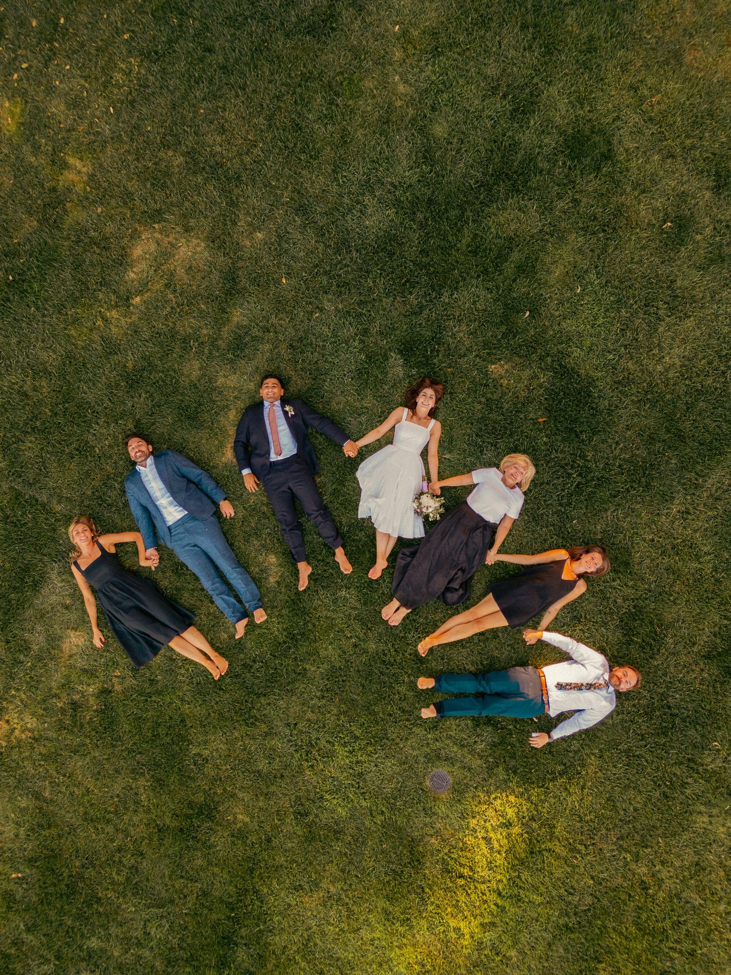 Aerial drone view of wedding party lying on green grass in a circle formation, bride in white dress at center.