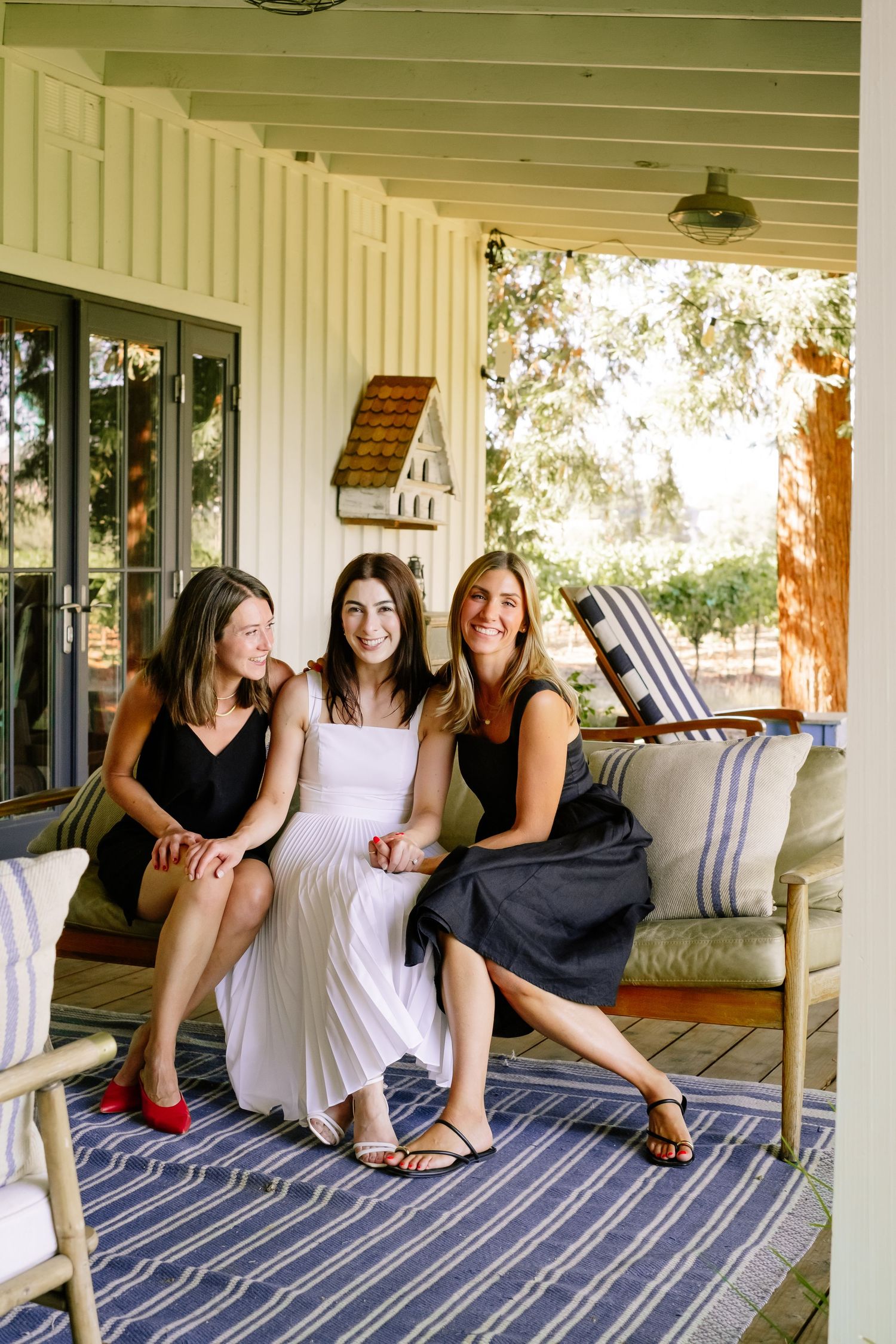 Bride in white dress sits with two women in black dresses on a porch swing outdoors.