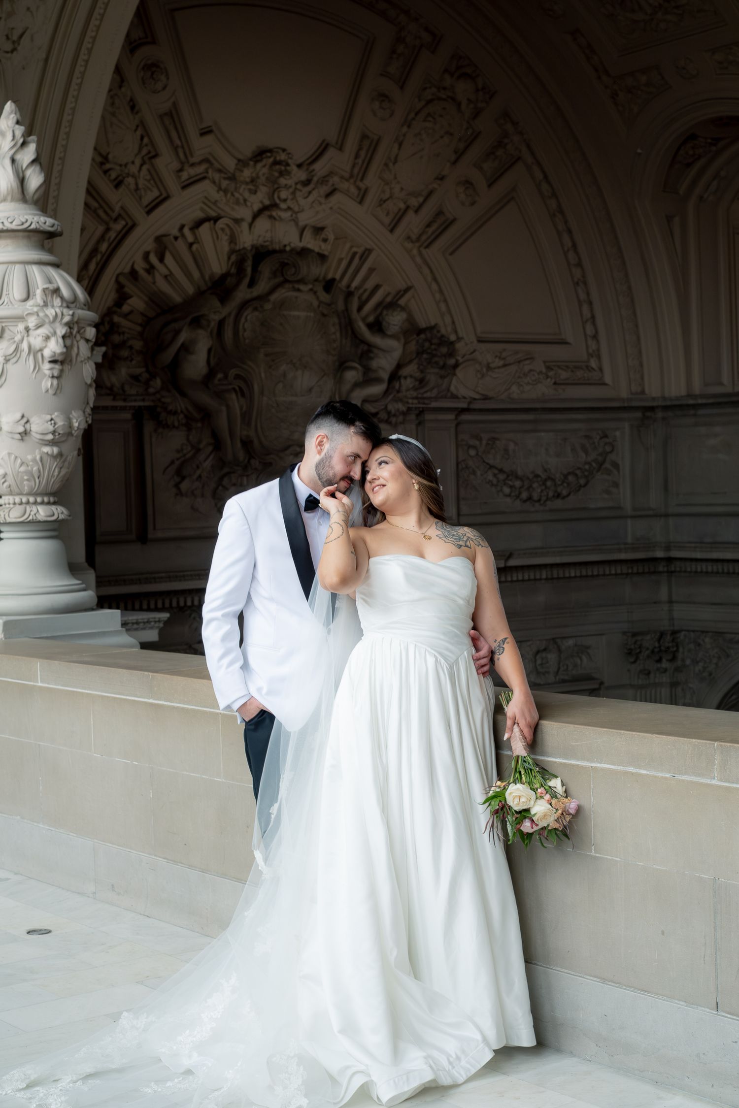Bride and groom pose together in front of ornate stone architecture, she holds a bouquet in a strapless white gown.