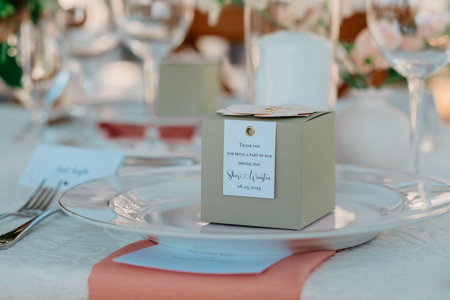 An elegant wedding favor box with a personalized label sits on a white plate at a beautifully set reception table.
