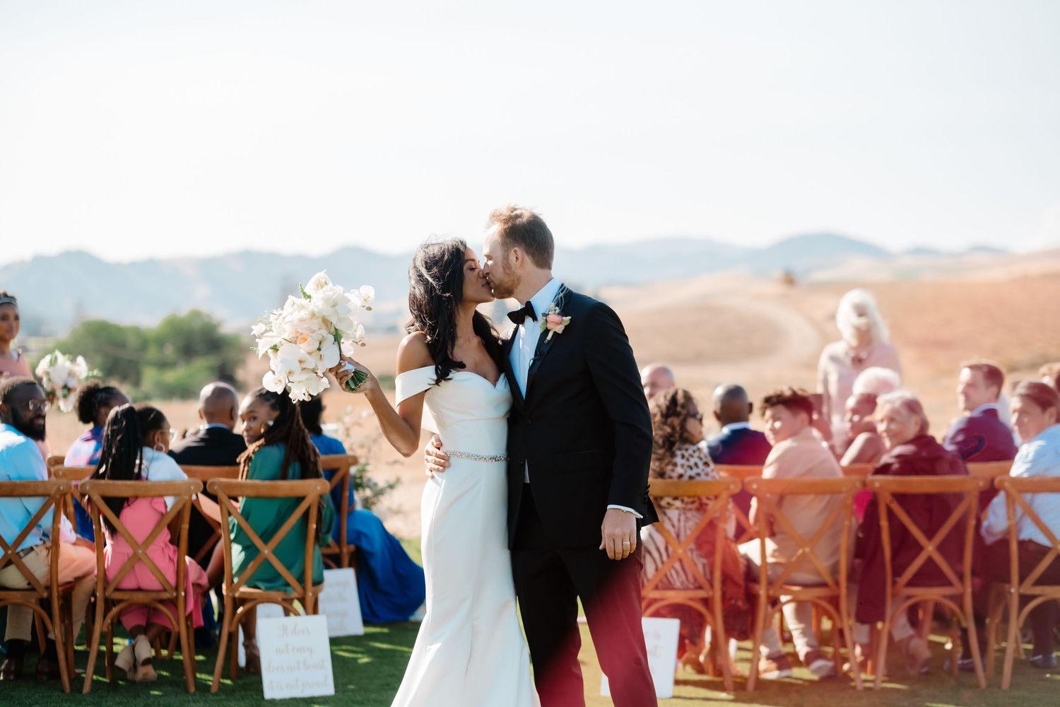 Newlyweds share first kiss at outdoor wedding ceremony with guests seated in colorful chairs.