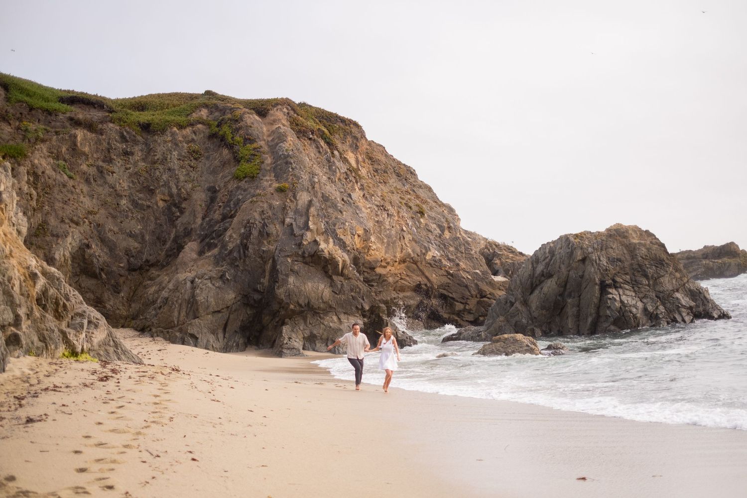 Engaged couple walking hand-in-hand along secluded sandy beach surrounded by dramatic rocky coastal cliffs.