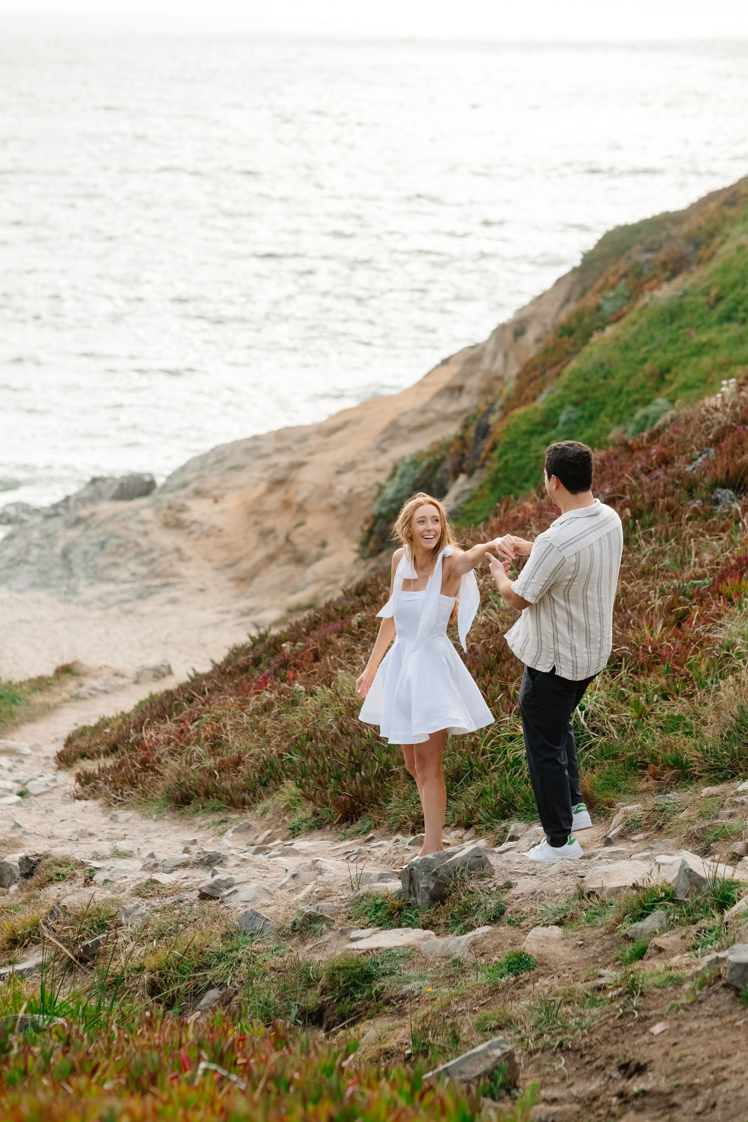 Couple holding hands on a rocky coastal cliff path with ocean waves in the background.