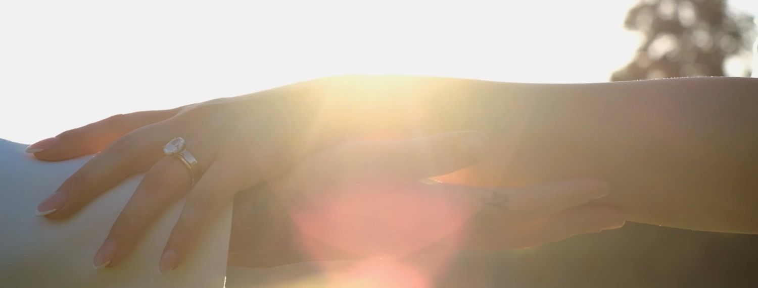 Close-up of bride and groom holding hands in golden sunlight, showcasing a sparkling engagement ring on the bride's finger.