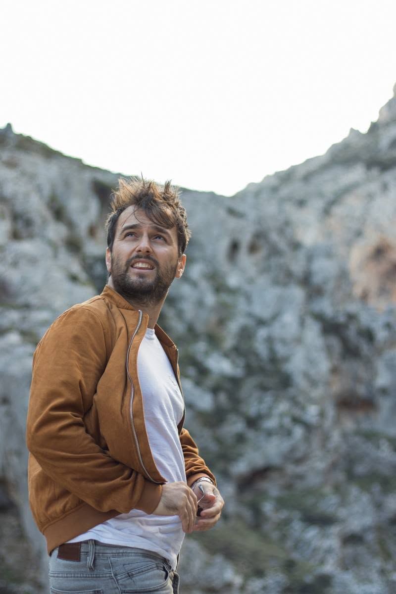 Bearded man in brown jacket looks up while standing against rocky mountain backdrop outdoors.