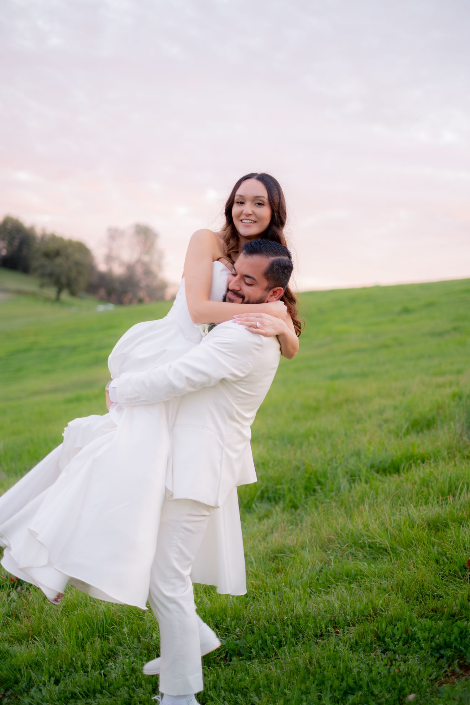 Groom in white suit lifting smiling bride in white dress on a green field at sunset.
