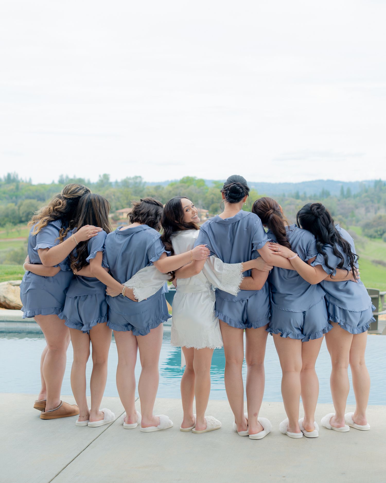 Bride and bridesmaids in matching blue sets embrace by a pool with scenic countryside views.