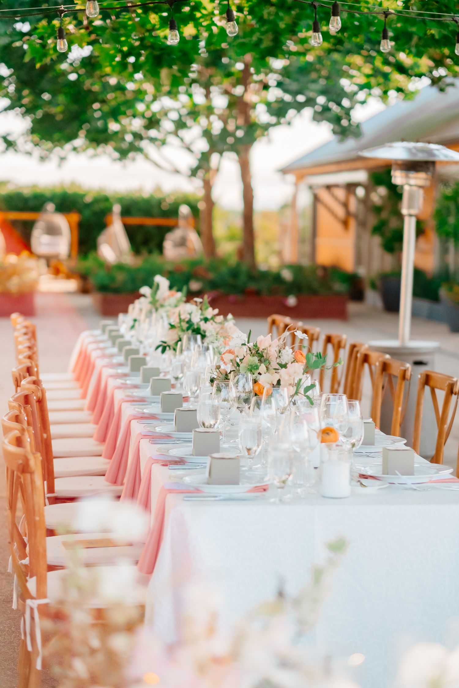 Elegant outdoor wedding reception table with pink linens, floral centerpieces, and string lights.