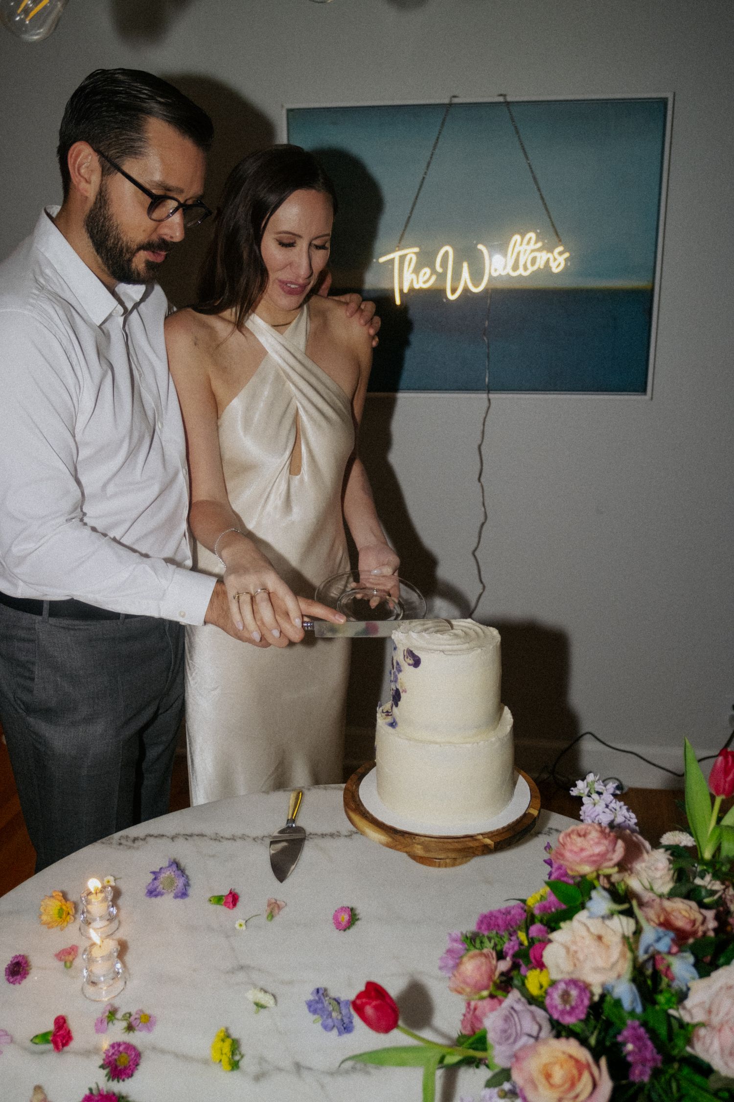 Newlyweds cutting their white two-tier wedding cake beneath a neon sign reading The Waltons, surrounded by colorful flowers.