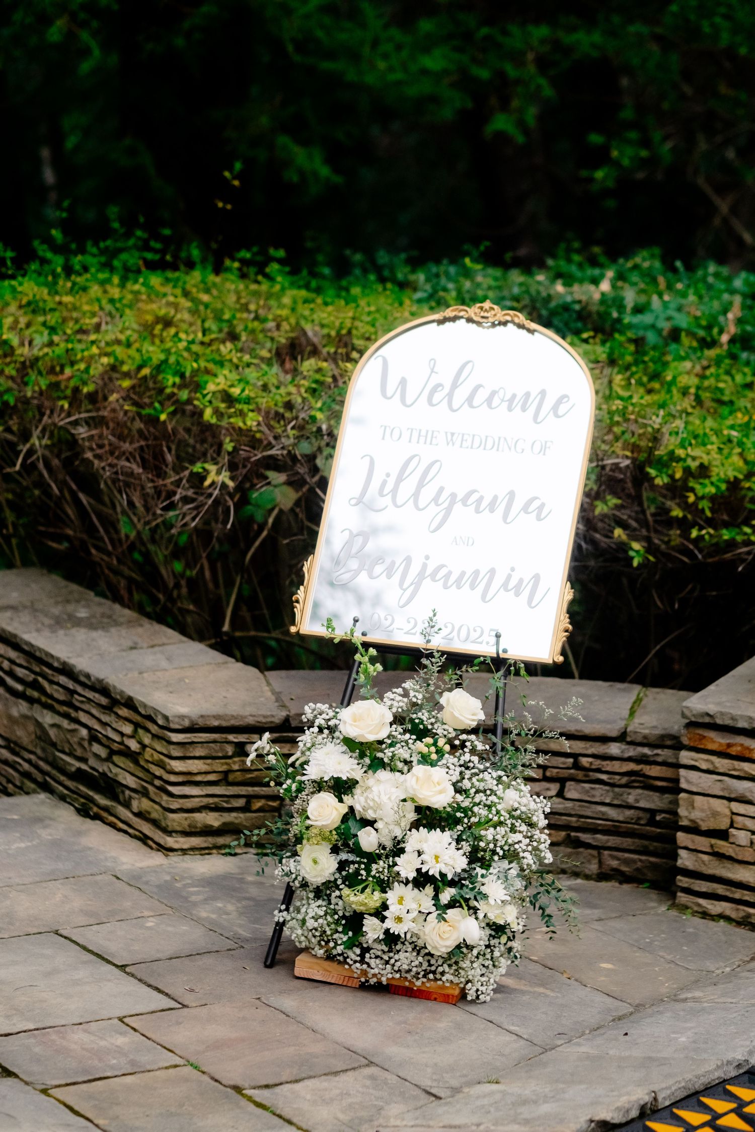 Welcome sign for Lillyana and Benjamin wedding with white floral arrangement on stone steps.