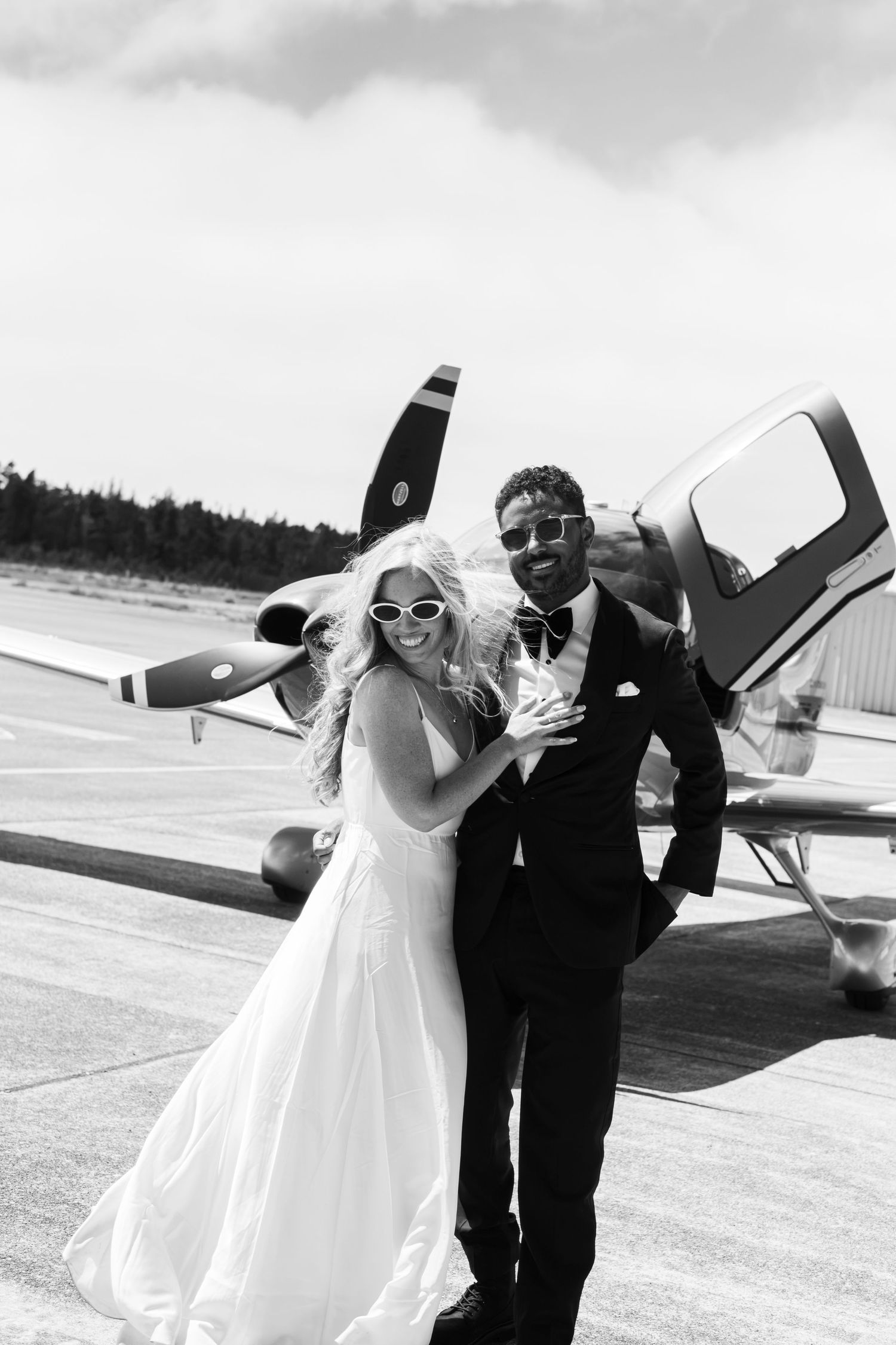 Black and white photo of a bride and groom posing in front of a small propeller plane on a runway.