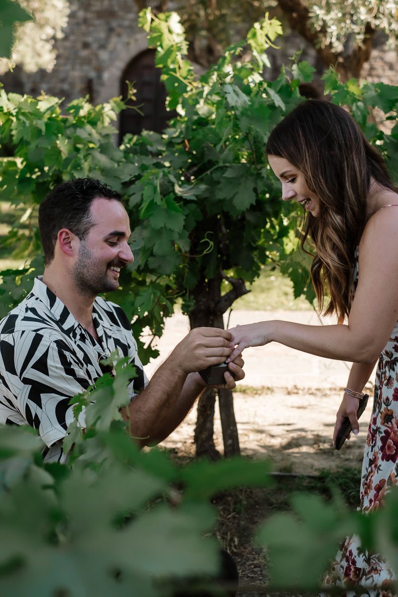 Smiling man proposing with ring to happy woman leaning forward in a lush vineyard.