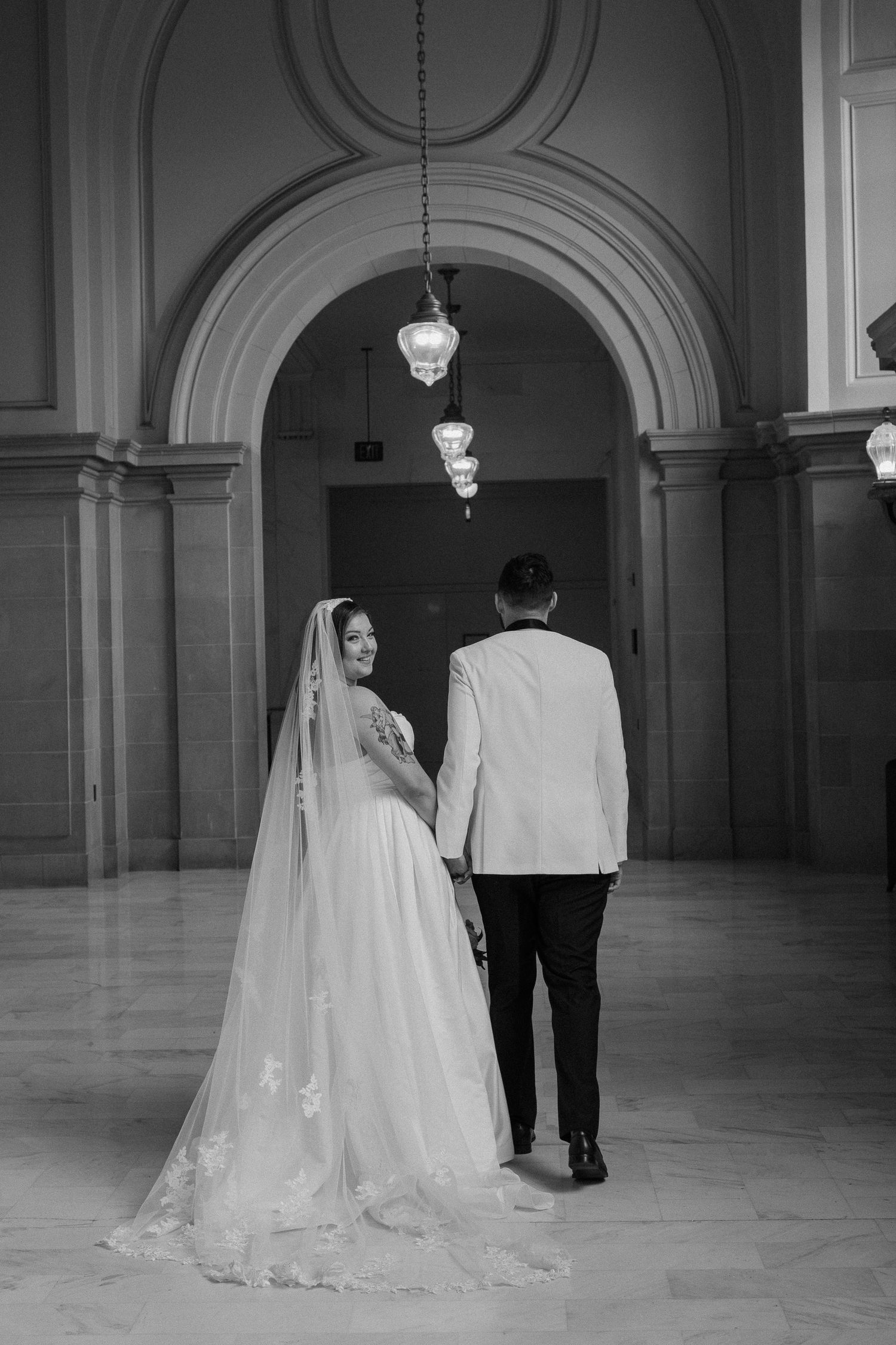 Black and white photo of a bride and groom walking hand in hand through an elegant arched hallway.