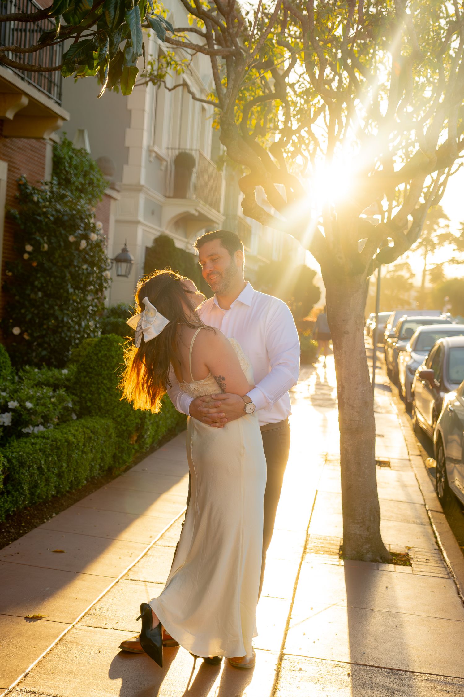 Couple embracing on sunlit sidewalk at golden hour, woman in cream slip dress and man in white shirt.
