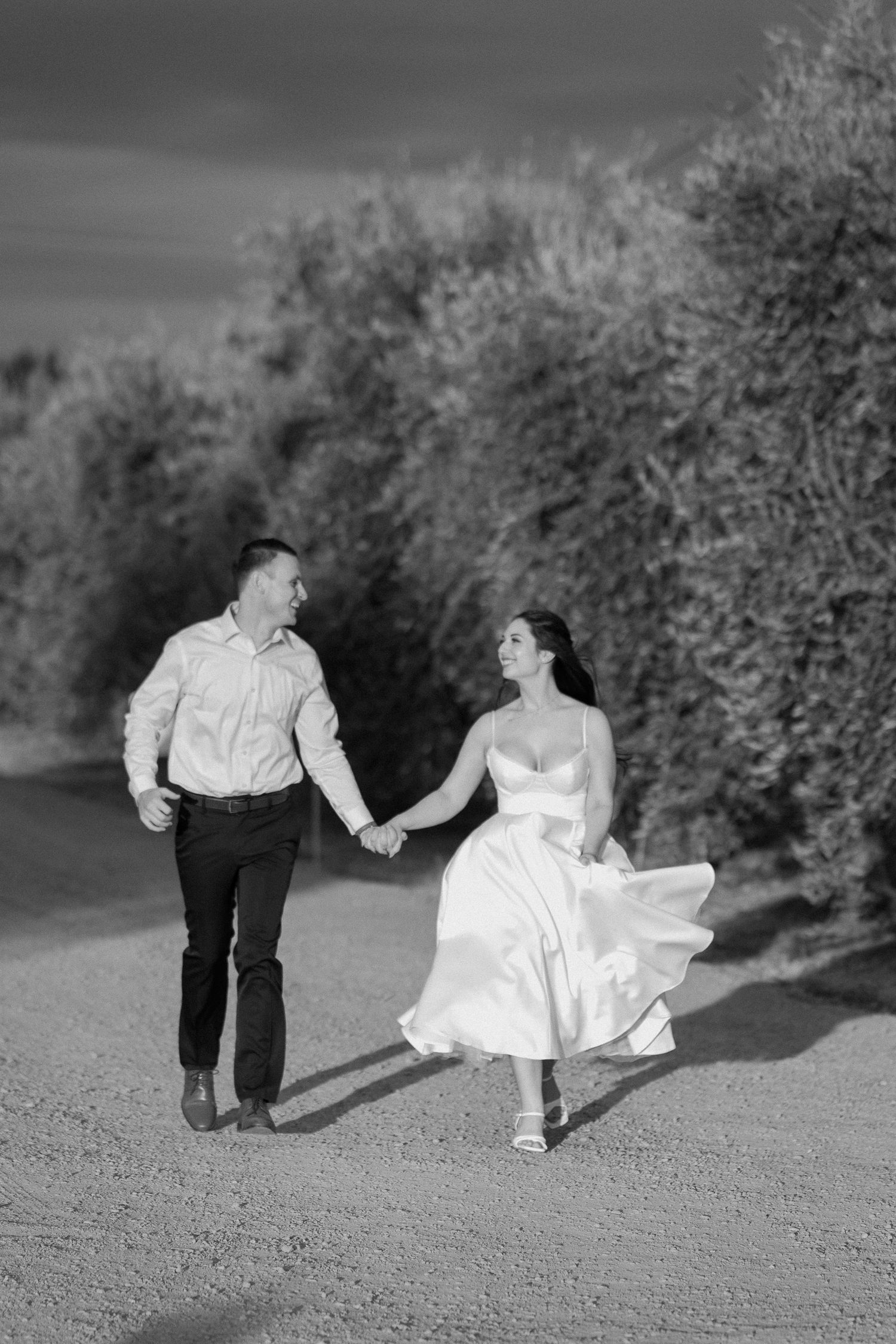 Black and white photo of a couple holding hands and walking joyfully down a sunlit path lined with olive trees.