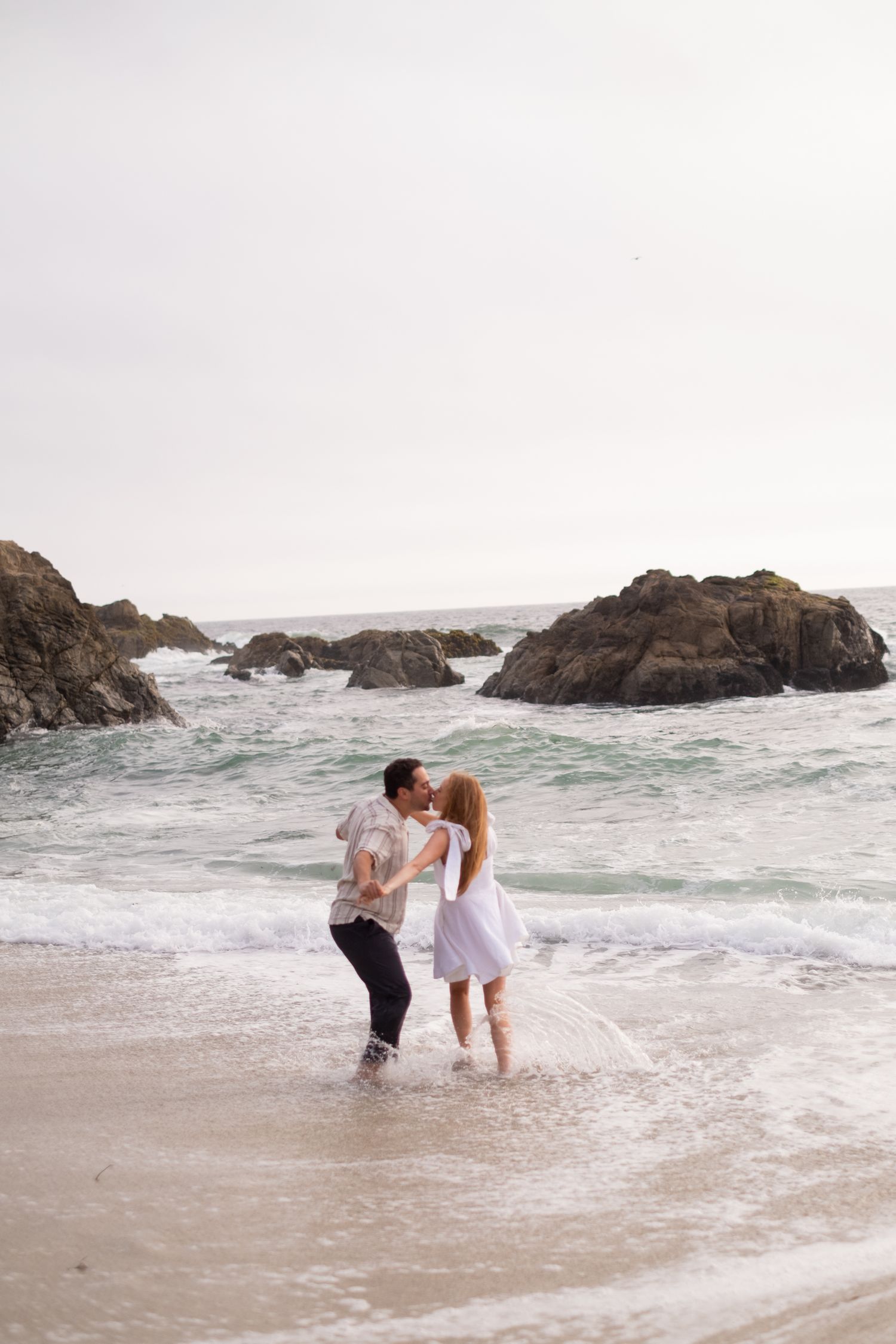 Couple kissing in shallow ocean waves on a sandy beach with rocky outcroppings in background.