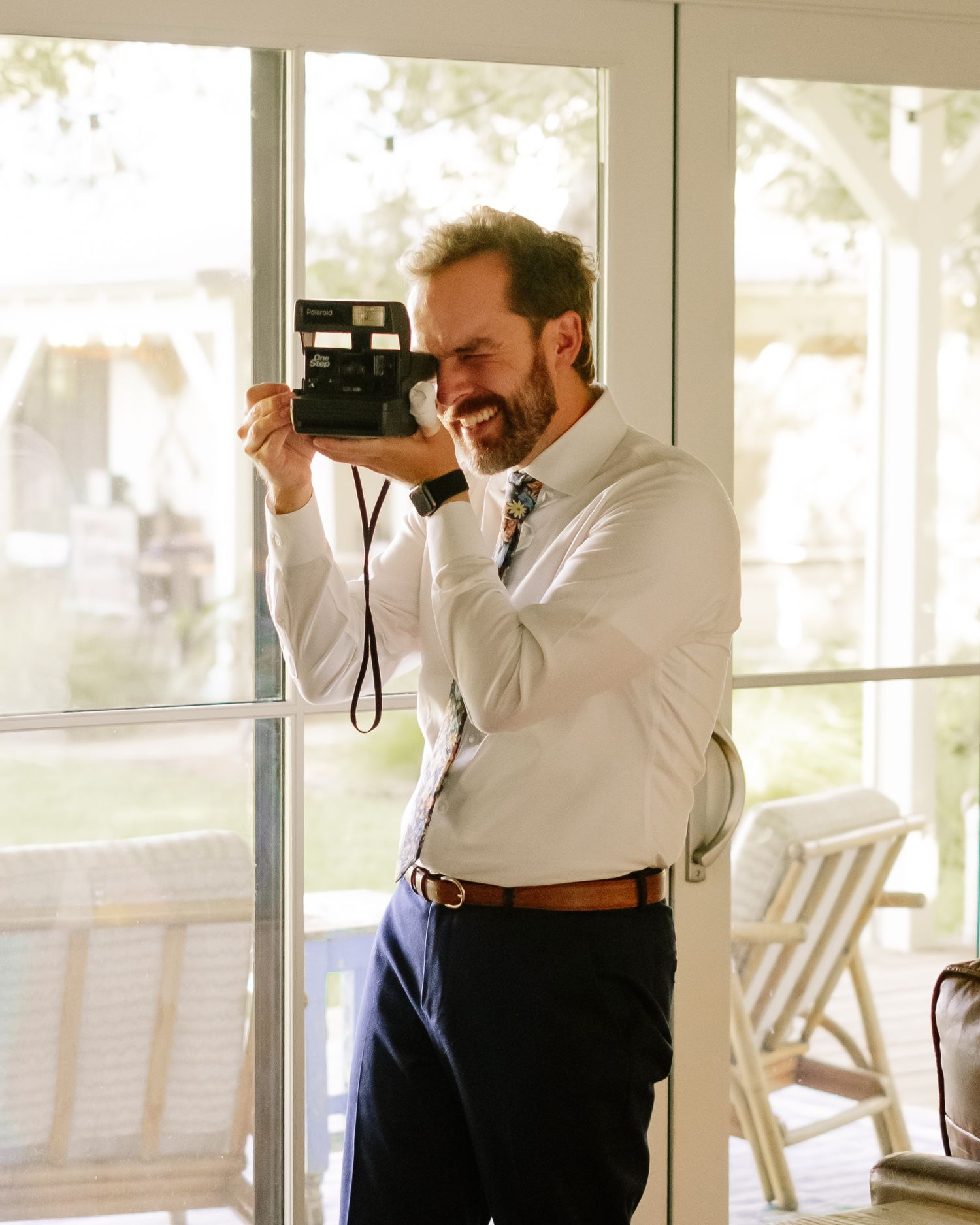 A smiling man in a white shirt and navy pants holds a Polaroid camera up to take a photo indoors.