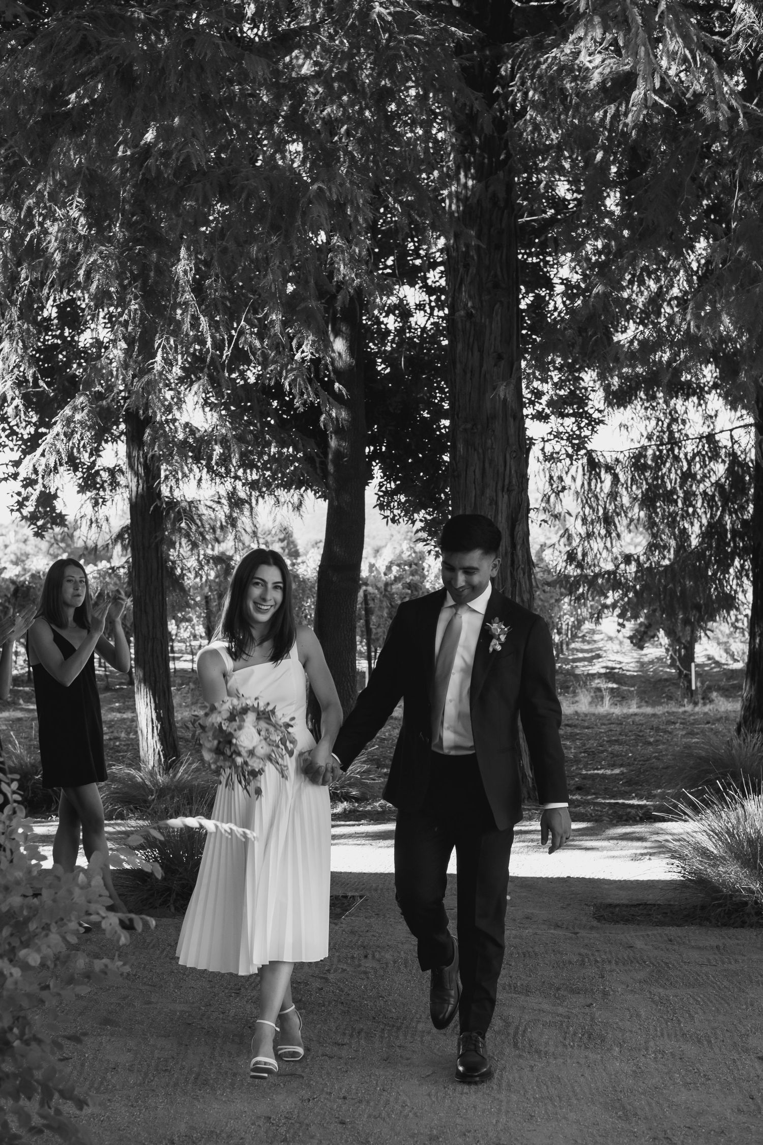 Black and white photo of bride and groom walking hand in hand through tree-lined path.