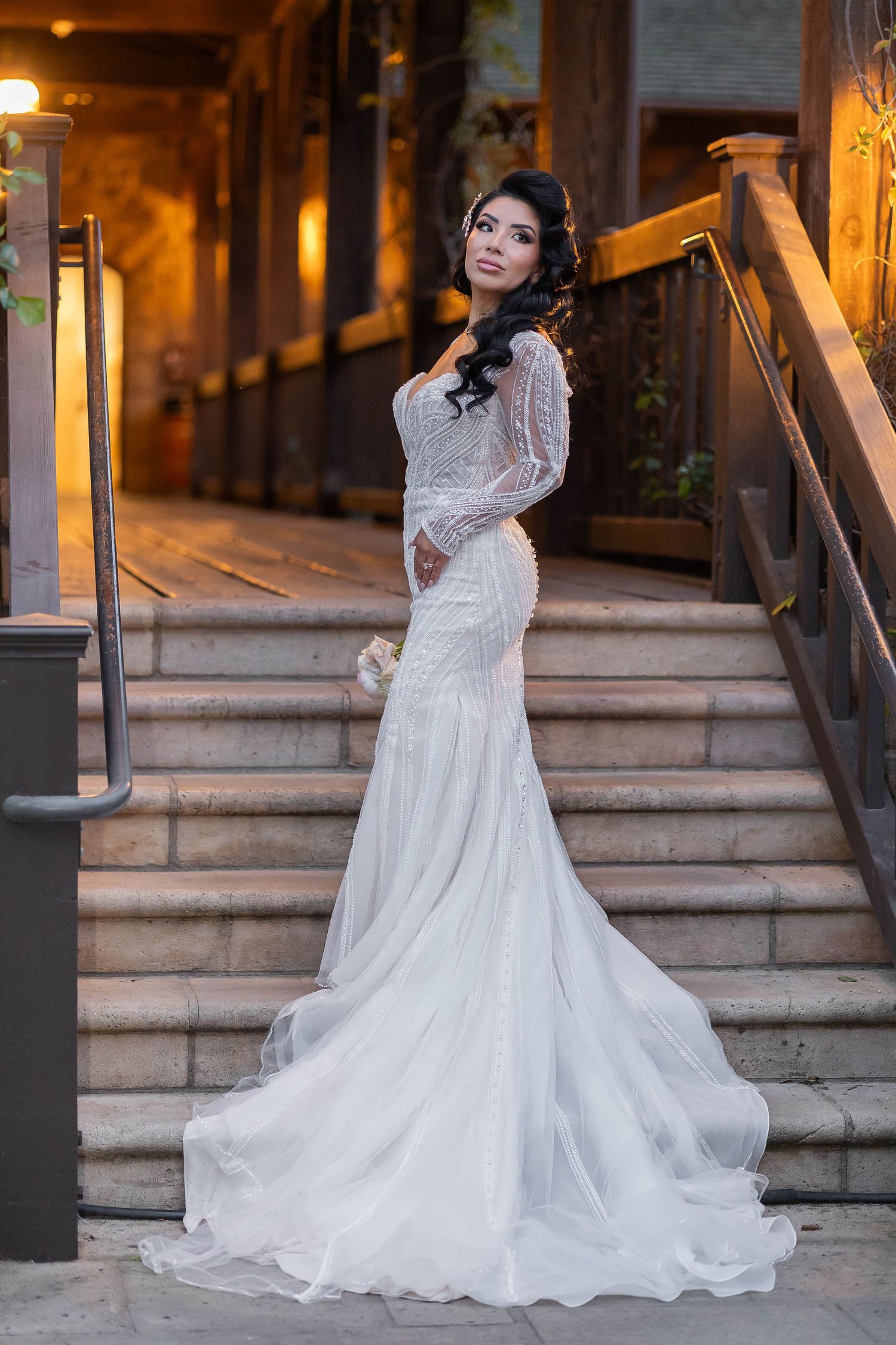 Elegant bride in a long-sleeve beaded mermaid gown posing on stone steps at night.