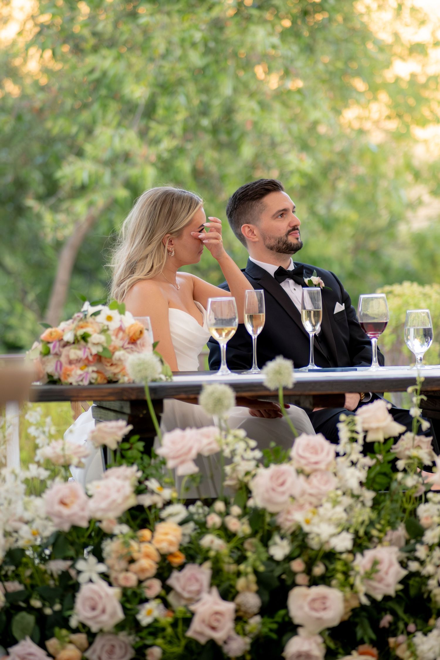 Emotional bride wiping tears at sweetheart table surrounded by lush floral arrangements during wedding reception.