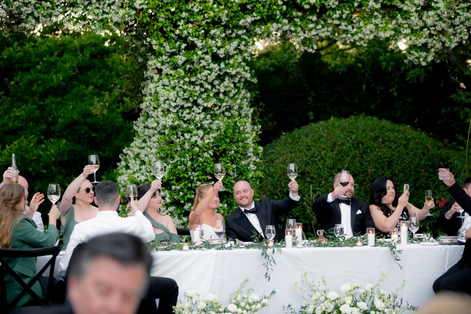 Wedding party raising champagne glasses in a toast at elegant outdoor reception with floral arrangements.
