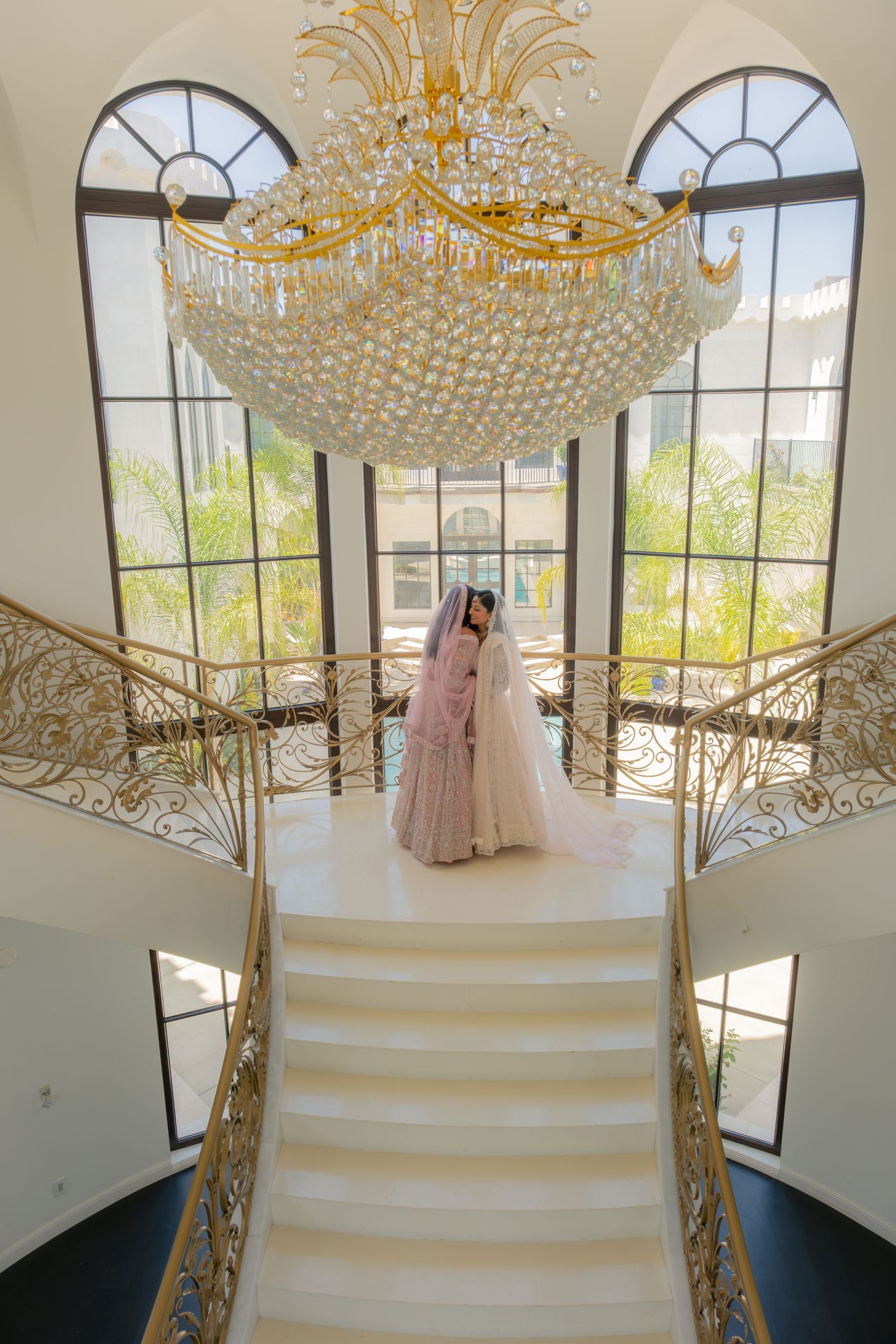 Bride in pink lehenga stands under grand crystal chandelier on elegant white staircase in luxury venue.